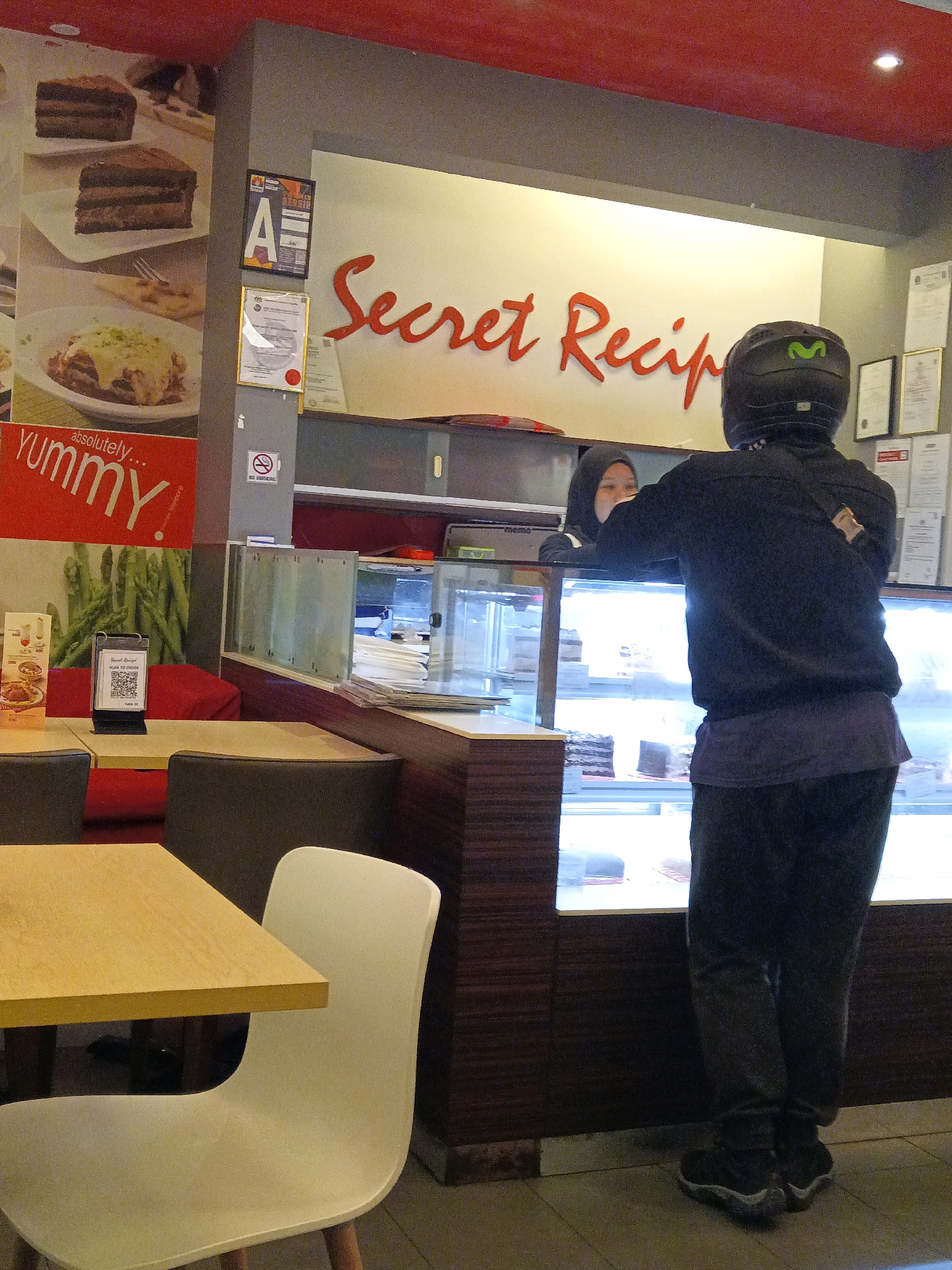 The image depicts the interior of a "Secret Recipe" cafe in Sepang, Malaysia. In the foreground, a light wood table and a white chair are visible. Further back, a person wearing a black helmet with a green "M" logo and dark clothing is standing at a counter, facing away from the camera. This person appears to be interacting with a staff member, a woman wearing a hijab, who is partially visible behind the counter. The counter features a glass display case containing cakes. Behind the counter, a large red sign reads "Secret Recipe." The walls are painted white and red, and there are various certifications and decorative items displayed on the wall. A "No Smoking" sign is also present. The overall environment suggests a commercial establishment serving food and beverages.
