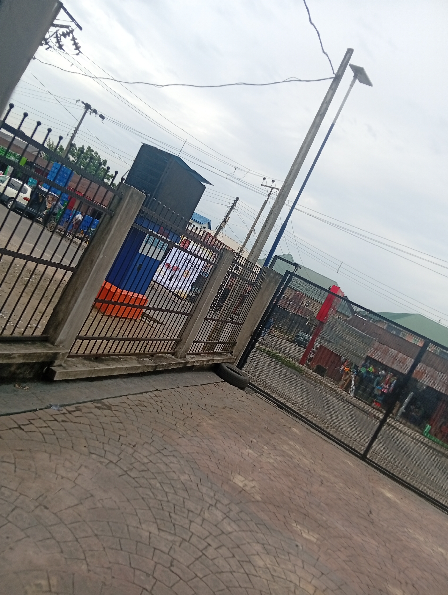 The image depicts an outdoor scene in Rumuepirikom, Nigeria. In the foreground, a textured paved walkway with a pattern of curving bricks is visible. A dark metal fence with vertical bars and concrete posts runs across the middle ground, separating the walkway from a street area. Beyond the fence, several structures are visible, including a dark blue shipping container and what appears to be a small kiosk or market stall with merchandise displayed. A white banner with text is attached to the fence. In the background, there are buildings with metal roofs, trees, and a street with a few vehicles, including a white car and a motorcycle with a rider. Overhead, power lines and utility poles are present against an overcast sky. The overall impression is that of a commercial or residential area within the city.