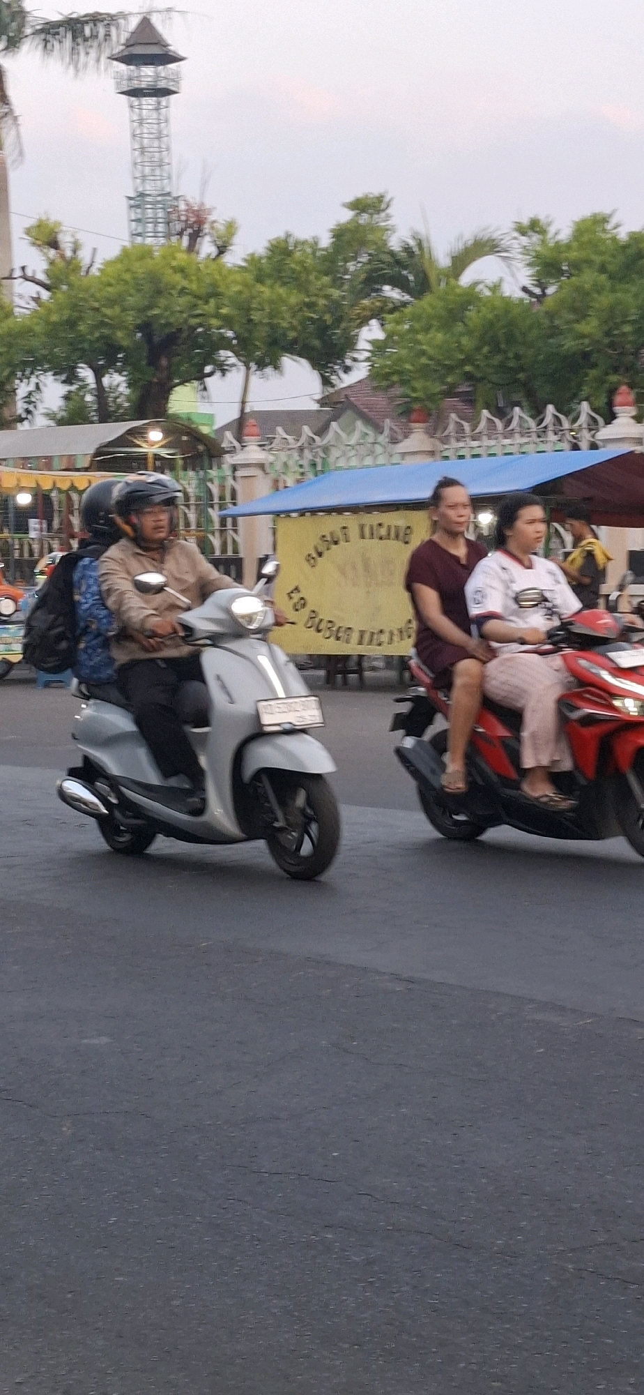 In Desa Donohudan, Indonesia, two motorbikes are visible on a paved road. The motorbike in the foreground is silver, with a rider wearing a light brown shirt and a black helmet. A passenger in a blue patterned shirt is seated behind the rider. The motorbike has a license plate that reads "AD 5902 BSF". To the right, a red motorbike carries two people: a woman in a dark maroon dress and a woman wearing a white shirt with red accents and pink patterned pants. Both are looking forward. Behind them, a yellow banner with red writing, possibly advertising "ES BUBUR KACANG", is displayed. In the background, trees with green foliage and a building with a decorative white fence are present. Further in the distance, a tall metal tower with a pointed roof stands against a pale sky. The overall lighting suggests it is either early morning or late afternoon.