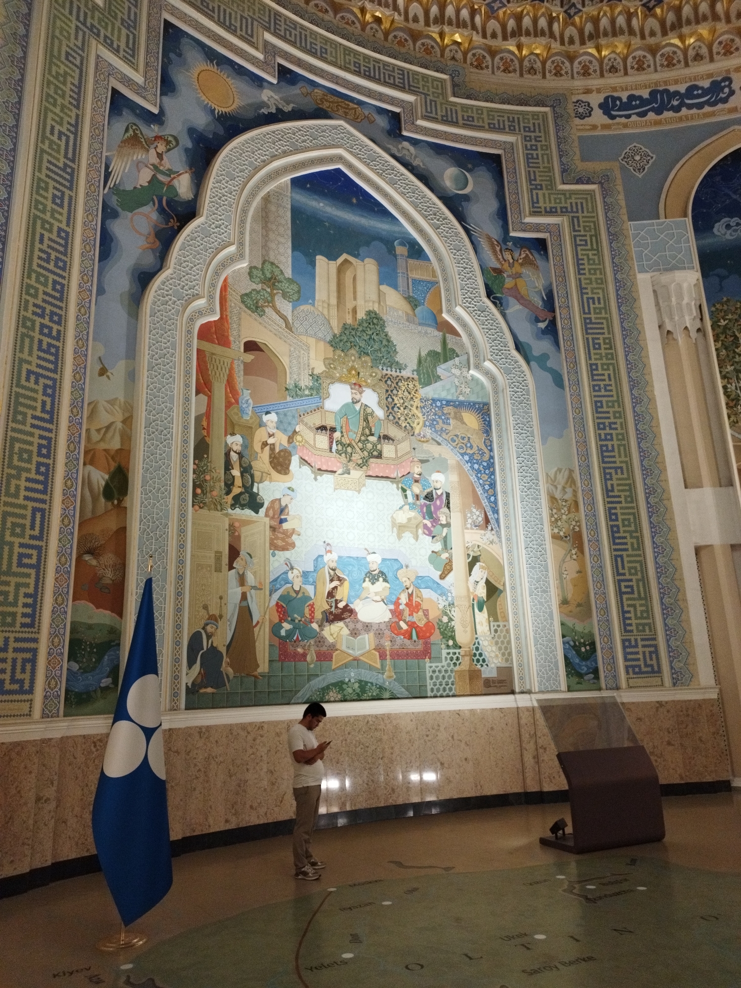 The image depicts a man standing and looking at his phone in an interior space with a large, ornate mural on the wall. The mural features intricate geometric patterns and a detailed painting depicting figures in traditional attire, architectural elements, and a celestial scene with a sun and moon. A blue flag with three white circles is positioned to the left of the man. The floor appears to be a map or a decorated surface with place names visible. The overall environment suggests a cultural or historical site, possibly a museum or a governmental building. The lighting is bright, with some areas illuminated by sunlight or artificial lights creating reflections on the floor.