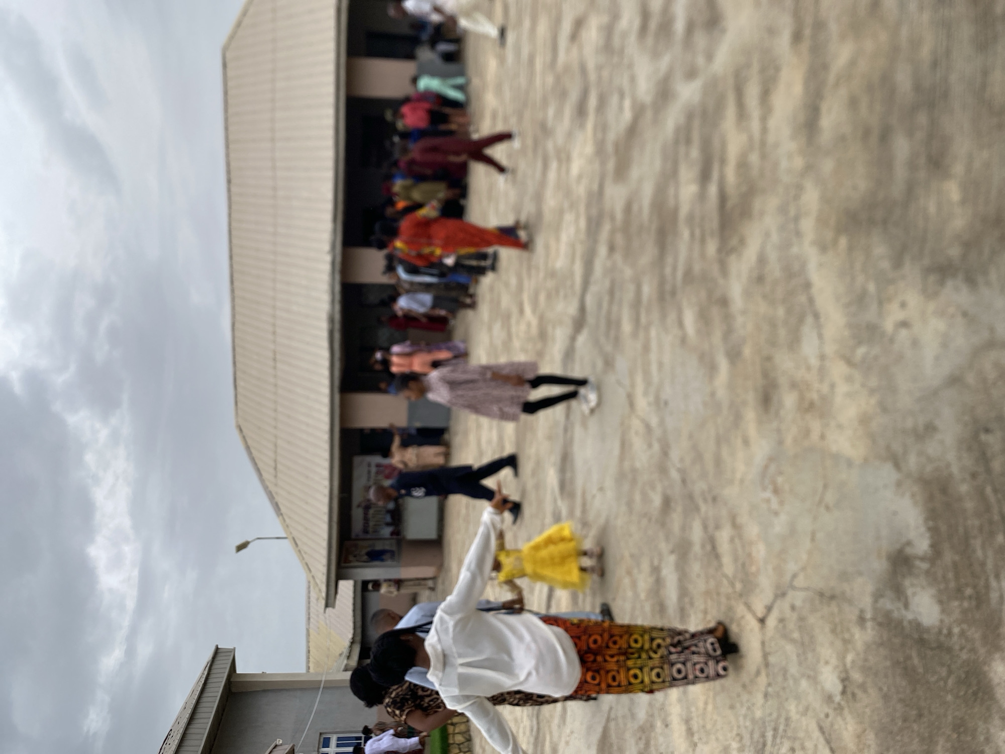 The image shows a group of people gathered in an outdoor space, possibly a courtyard, in Rukuba Road, Nigeria. The environment is characterized by a building with a light-colored facade and a light grey, overcast sky above. The ground is a textured, light-colored concrete.
The people in the image vary in age and attire. Some are wearing casual clothing, while others are dressed in more formal attire, including colorful, patterned fabrics. Some individuals appear to be walking or moving, while others are standing. A child wearing a yellow dress is visible.