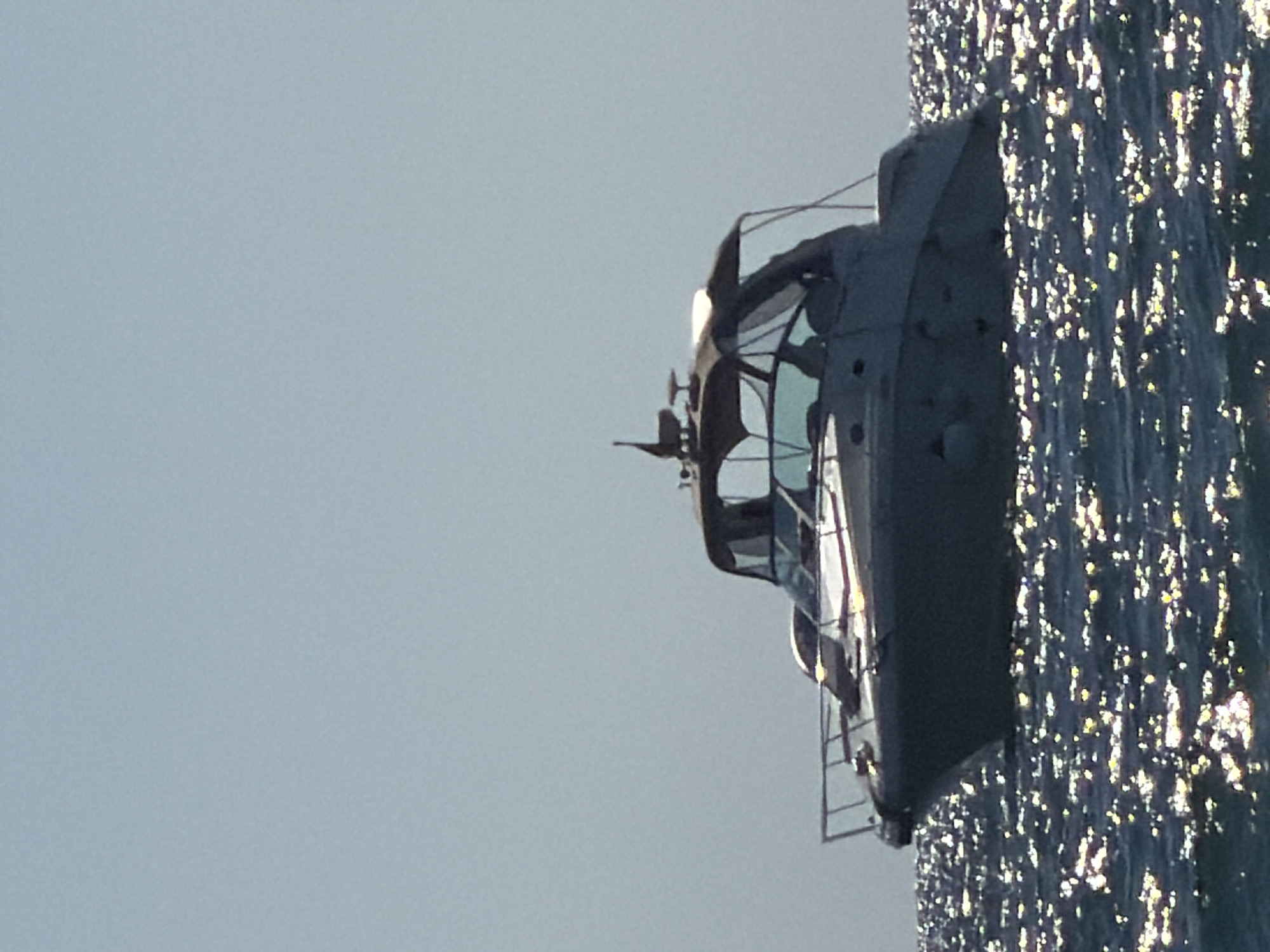 A medium-sized motorboat is positioned on the water, its hull reflecting the bright sunlight, creating a shimmering effect on the surface. The boat appears to be moving, with a slight incline suggested by the wake or its placement on the waves. The sky above is a pale blue with no visible clouds. The scene is likely outdoors, in a coastal environment, potentially the Black Sea, given the mention of Sochi, Russia. The image does not contain any people or specific landmarks to confirm its exact location within Sochi.