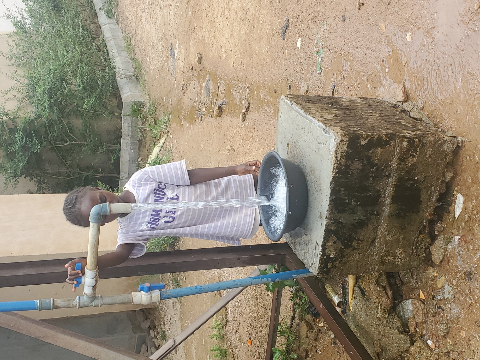 A young person stands outdoors at a water spigot. They are filling a black bowl with water. The spigot is constructed with blue PVC pipes. A concrete block sits next to the spigot, and the bowl rests on it. The area appears to be muddy. The location is likely Barakin Akawo, Nigeria.