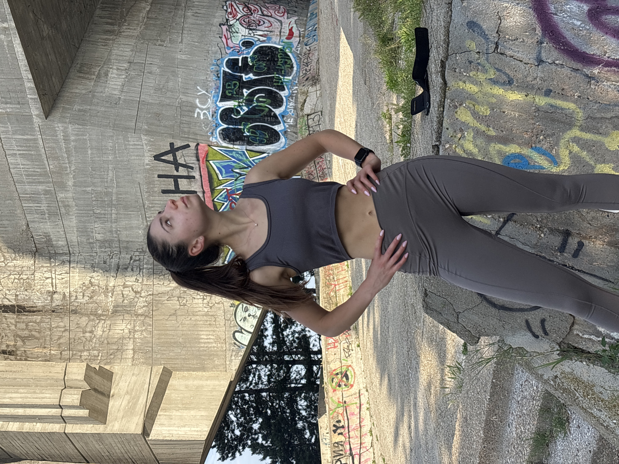 The image captures a young woman posing outdoors in what appears to be an urban or industrial setting, possibly under a concrete bridge or overpass in Varna, Bulgaria. She stands with her hands on her hips, head tilted upwards and to her left, looking away from the camera with a neutral expression. She is dressed in athletic wear, consisting of a dark grey ribbed crop top that reveals her midriff, and matching dark grey leggings. Her dark hair is pulled back in a ponytail, and she wears a black smartwatch on her left wrist. Her fingernails are painted light pink.
The background is dominated by large, weathered concrete structures, extensively covered in colorful graffiti. Visible tags and letters include "HA", "BCY", and other Cyrillic characters in black, blue, green, red, yellow, and purple spray paint. The ground is a mix of cracked concrete and dirt, with sparse patches of green grass. Natural daylight suggests it is daytime, likely with clear skies, casting some shadows. A black arm-strap object lies on the ground near the woman's right foot. The scene evokes an urban-athletic aesthetic, common in city fringe areas.