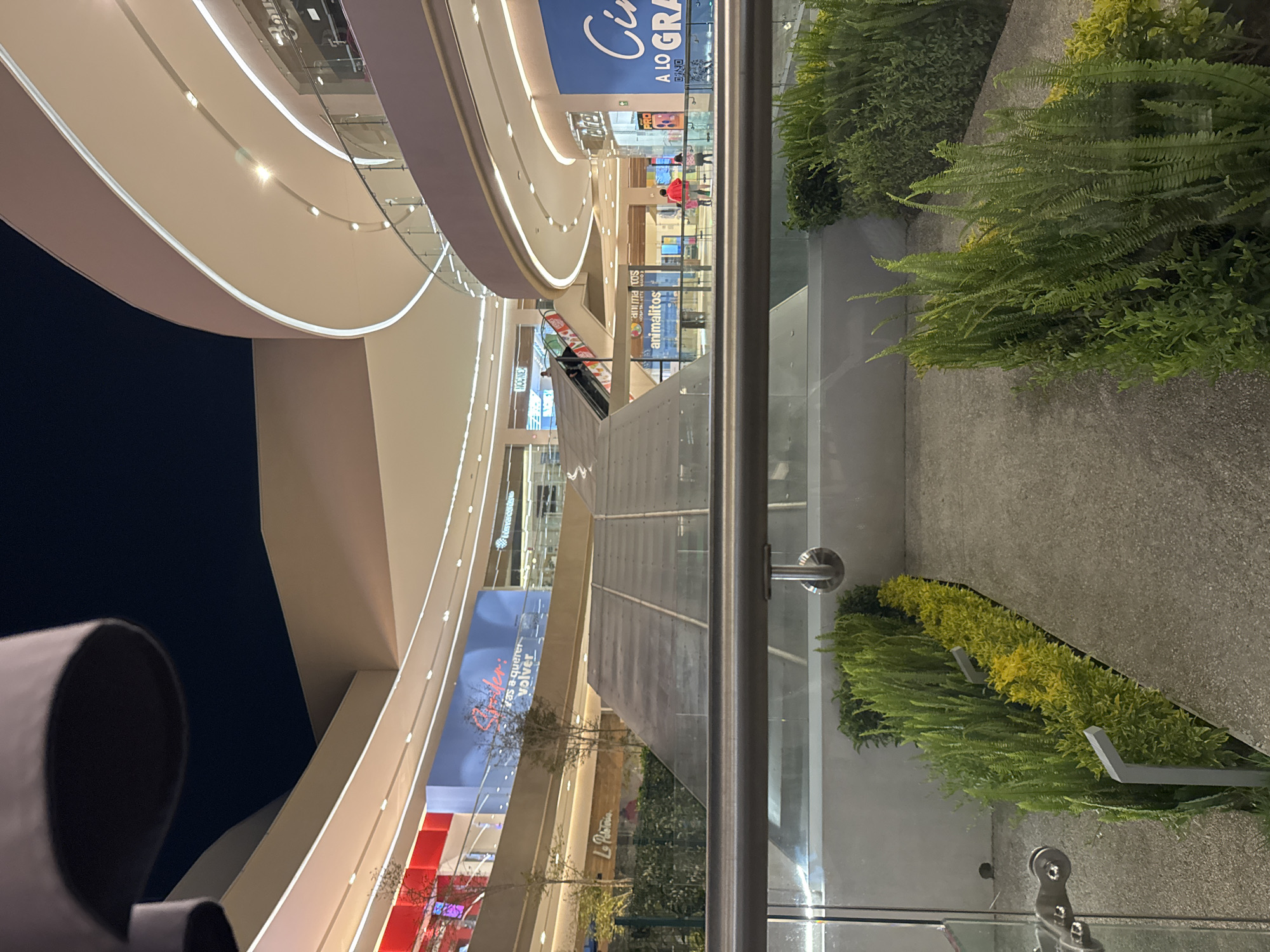 The image, taken at an angle, captures the interior of a modern shopping mall in San Bernardino Tlaxcalancingo, Mexico. The environment is brightly lit with curved, layered architectural elements dominating the scene. Large, curved white structures with embedded lighting create a sense of depth. Glass barriers line the walkways, offering views of greenery planted below. Escalators are visible, indicating multiple levels within the mall. Banners and signs advertising businesses are present, including recognizable names like "Cinemex" and "Animalitos." The color palette is neutral with the white architecture contrasting against the dark night sky visible through the upper levels. The presence of greenery and the clean, modern design contribute to the mall's ambiance.