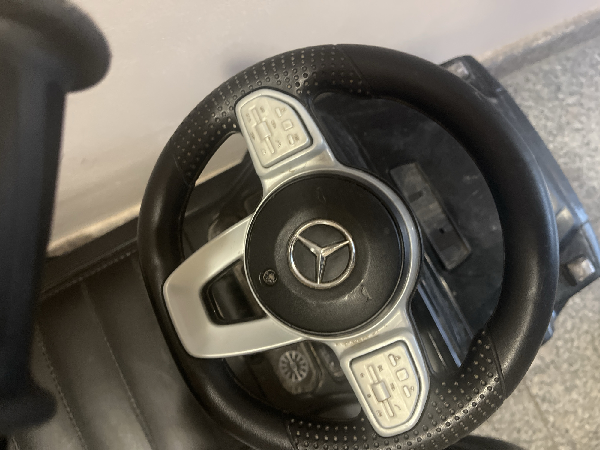 The image depicts a close-up, overhead view of a Mercedes-Benz steering wheel. The steering wheel is black with silver accents around the central Mercedes logo and on the spokes. The wheel has a textured grip. The steering wheel is likely part of a toy car. The environment is undefined. No people, subjects, actions, or interactions are visible. The image does not provide information to specify the location is in Plovdiv, Bulgaria.