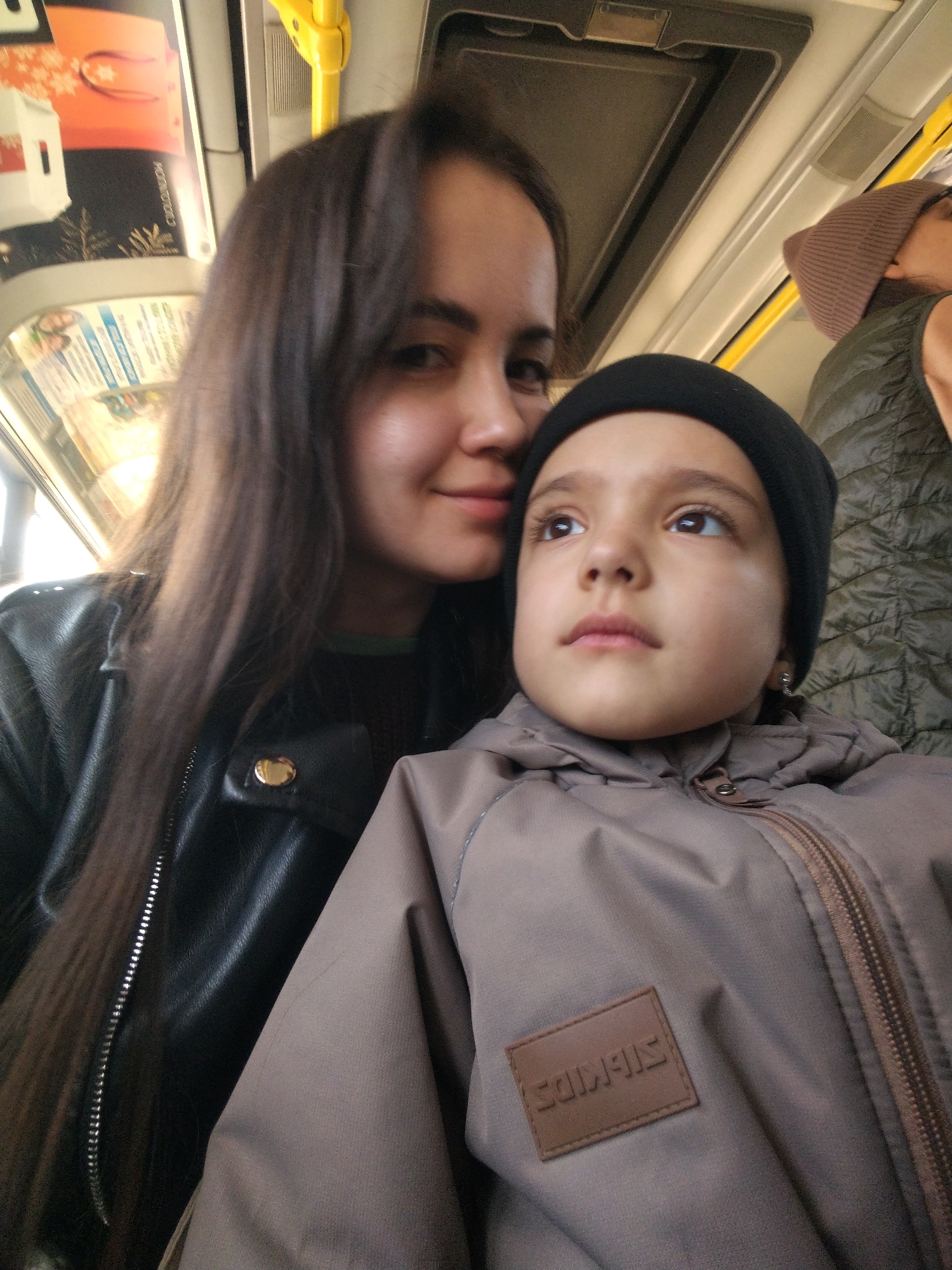 A woman and a child are in what appears to be public transportation, possibly a bus or a train. The woman has long, dark hair and is wearing a black leather jacket. She is looking at the camera. The child is wearing a black beanie and a gray jacket with a zipper. The jacket has a patch that reads "SIB Kids". The child is looking upward and to the left, away from the camera. The background suggests the interior of a vehicle with a window and overhead fixtures. Another person, wearing a brown beanie and a green puffy vest, is slightly visible behind them. The city is Chaikovsky, Russia.