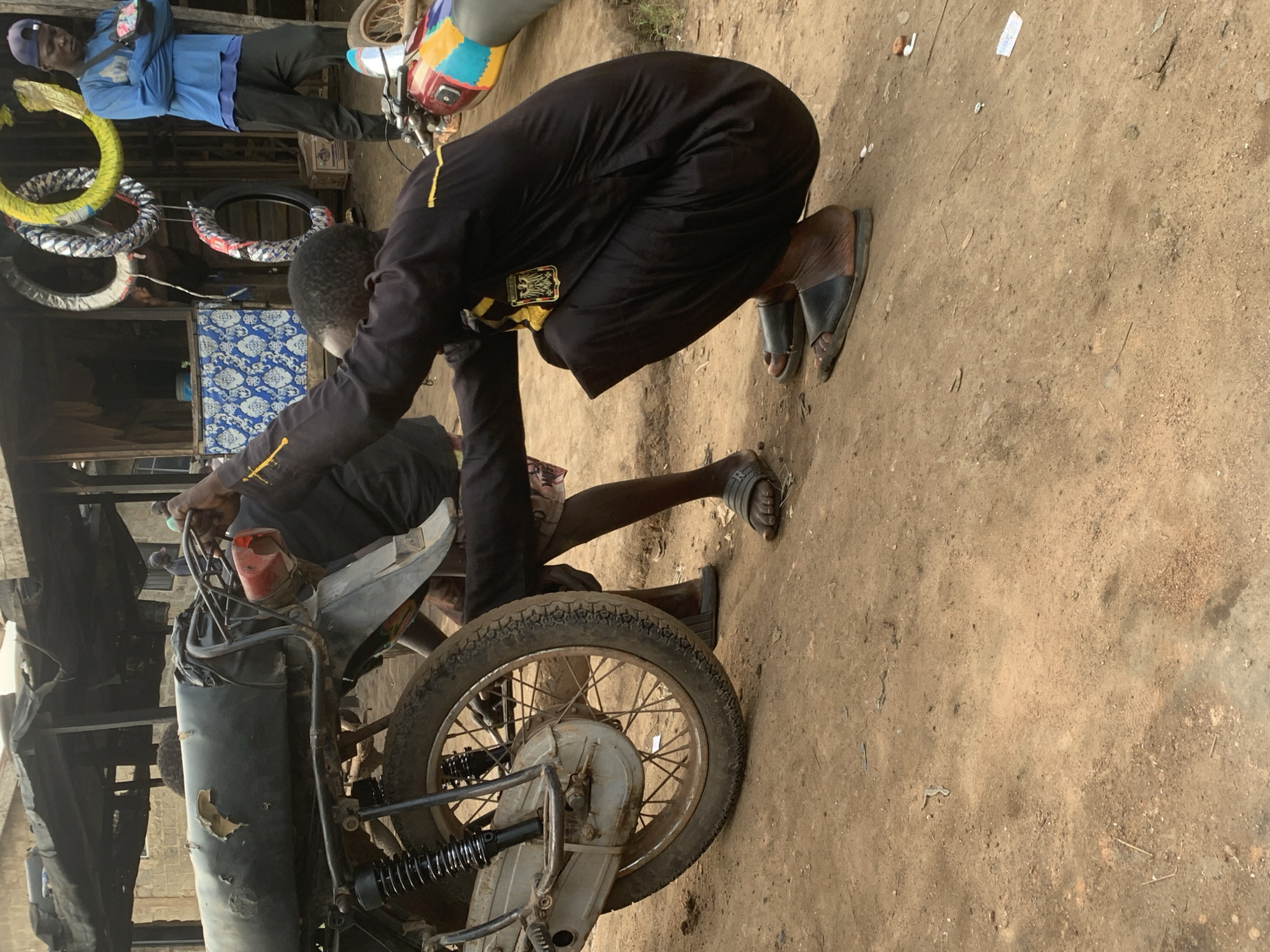 In the city of Shagamu, Nigeria, a man wearing a black robe with yellow trim and black sandals is crouched down, working on the rear wheel of a black motorcycle. The motorcycle has a worn, torn seat. Another person, mostly out of frame, is visible behind the motorcycle. In the background, a market stall is set up with various items, including several colorful motorcycle tires hanging from the ceiling. Another man in a blue long-sleeved shirt and a blue baseball cap stands to the right, looking down. The ground is composed of dry, sandy dirt.
