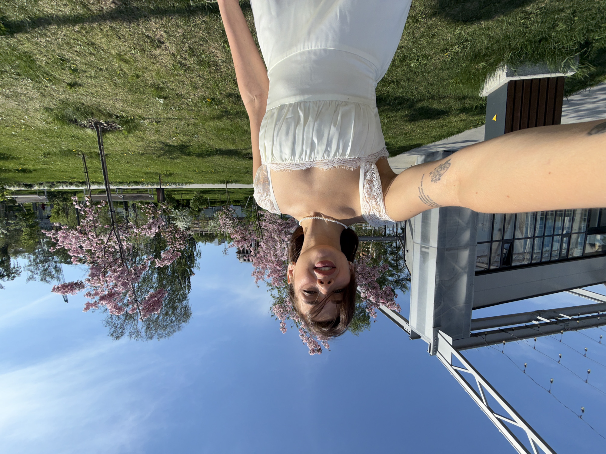 A young woman is posing for a selfie against a backdrop of trees with pink blossoms, blue sky, and modern architecture. She is wearing a white dress with lace detailing around the neckline and a pearl necklace. Her right arm is extended towards the camera, revealing a tattoo on her forearm. The location is Perm, Russia. In the background, there is a view of a metal structure, possibly part of a bridge or architectural element, against the blue sky. Trees with pink blossoms are visible on the left side of the image and modern architecture on the right.