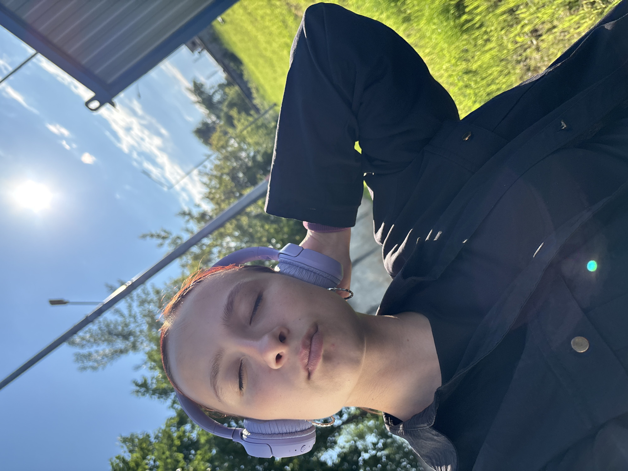 A young person, likely a woman or teenager, is depicted lying on their side outdoors, looking upwards towards the sky with their eyes closed. They are wearing light purple over-ear headphones and a dark, possibly black, jacket or top. A subtle, light-colored bracelet or watch is visible on their left wrist. Their expression is relaxed and serene, suggesting peace or contemplation.
The setting is an outdoor public space under bright daylight. The sun, a strong source of light, is positioned in the upper left, casting a noticeable glare and illuminating the subject's face while creating shadows on their clothing. The sky above is clear blue with some scattered white clouds. In the background, there is vibrant green grass or foliage. Parts of an outdoor structure, possibly a bus stop or shelter, are visible, featuring a blue or grey roof and transparent panels that reflect the sky and clouds. A streetlight pole is also present in the upper background. The overall impression is a sunny day in an urban or suburban environment. There is no visible text in the image.