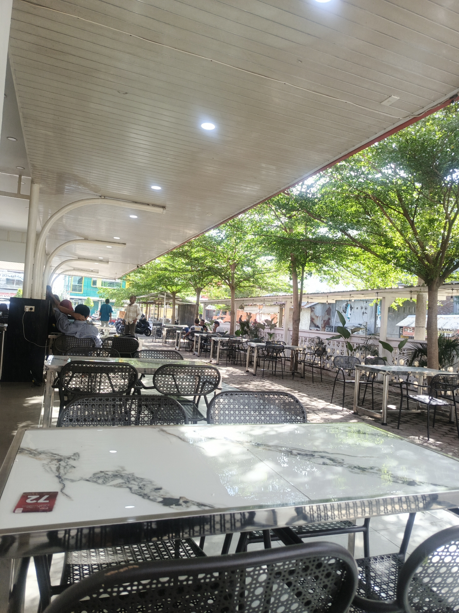 The image depicts an outdoor or semi-outdoor dining area, likely a cafe or restaurant, located in the city of Pantegajah, Indonesia. The scene is illuminated by bright daylight, with sunlight filtering through trees and casting shadows.
In the immediate foreground, multiple empty tables with glossy white marble-patterned tops and reflective chrome-finish frames are visible. Dark-colored chairs with woven, rattan-style backs and seats are positioned around these tables. A red square card marked "72" rests on the leftmost table. The table surfaces reflect portions of the sky and tree canopy.
The dining area extends under a flat, slatted white ceiling supported by white arched and straight columns. Recessed circular downlights and linear fluorescent light fixtures are integrated into the ceiling. Several individuals are present: two men are seated at a table on the left, one wearing a light blue shirt and a red cap, facing away from the camera. Further back, a man in a plaid shirt stands near a parked motorcycle. Other patrons are sparsely distributed at tables further into the covered area. A dark, rectangular speaker or equipment box is positioned on the far left, adjacent to a column.
Beyond the covered section, an open-air seating area is shaded by a line of mature trees with lush green foliage and slender trunks. More tables and chairs are arranged beneath these trees on a paved surface that displays dappled sunlight and shadows. A low, white-painted wall with decorative ventilation blocks separates this area from the background.
The background reveals an urban environment. Behind the wall, various structures are visible, including corrugated metal sheds and utilitarian buildings, some with laundry hanging. Further back, multi-story buildings with glass windows and varied architectural styles are present on the left, while more modest, possibly residential or commercial structures are on the right.
