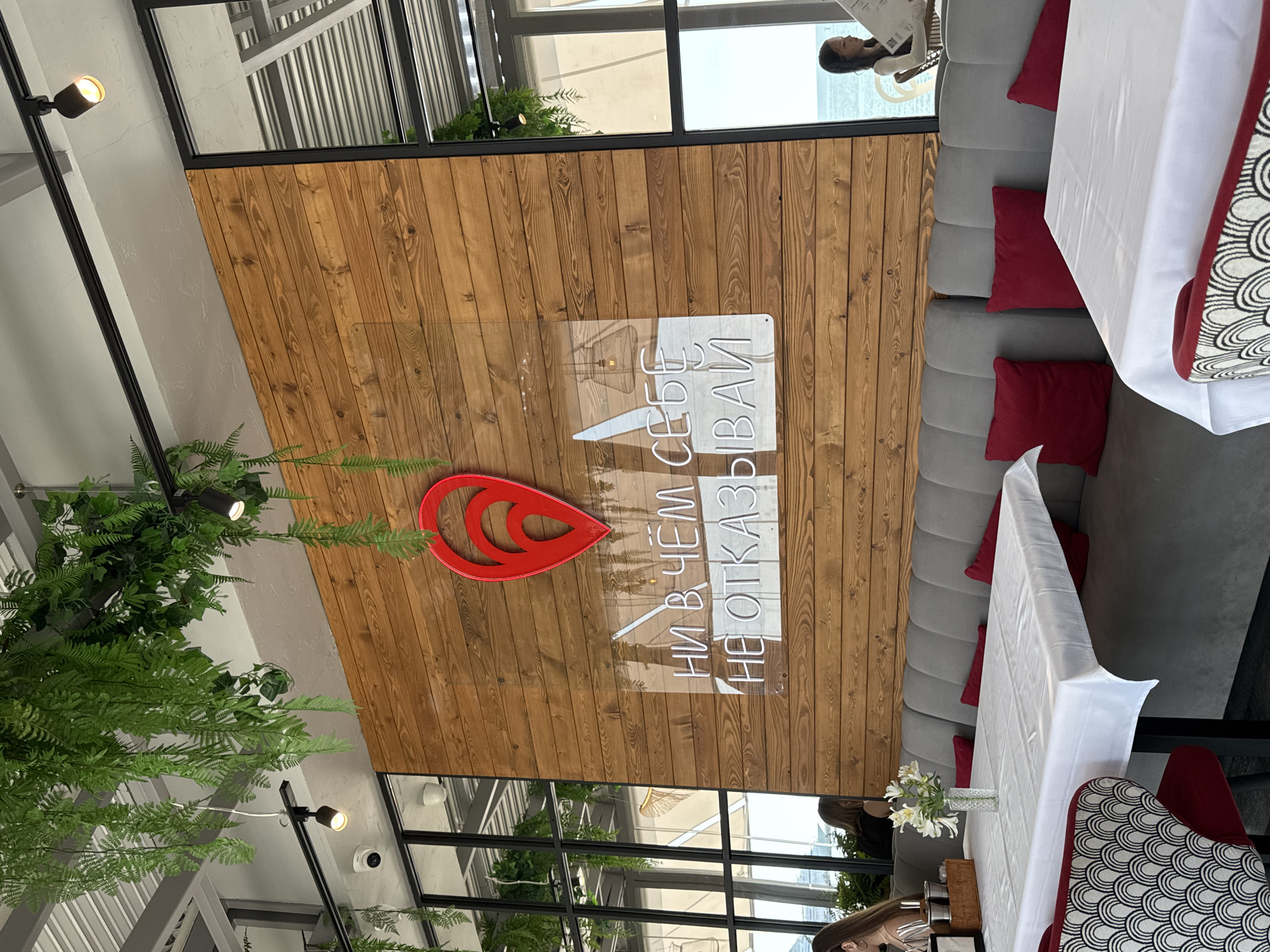 The image depicts the interior of a restaurant with a view of the sea. The restaurant features a wooden accent wall with a large red pin icon and a neon sign that reads "ни в чём себе не отказывай" (don't deny yourself anything). Several tables are set with white tablecloths and red cushions are placed on the banquette seating. Green artificial plants hang from the ceiling, and track lighting is visible. Through a large window or glass partition, the sea and a woman reading a menu can be seen. The scene is likely in Sochi, Russia, given the mention of a restaurant with a view of the sea and the Russian text.