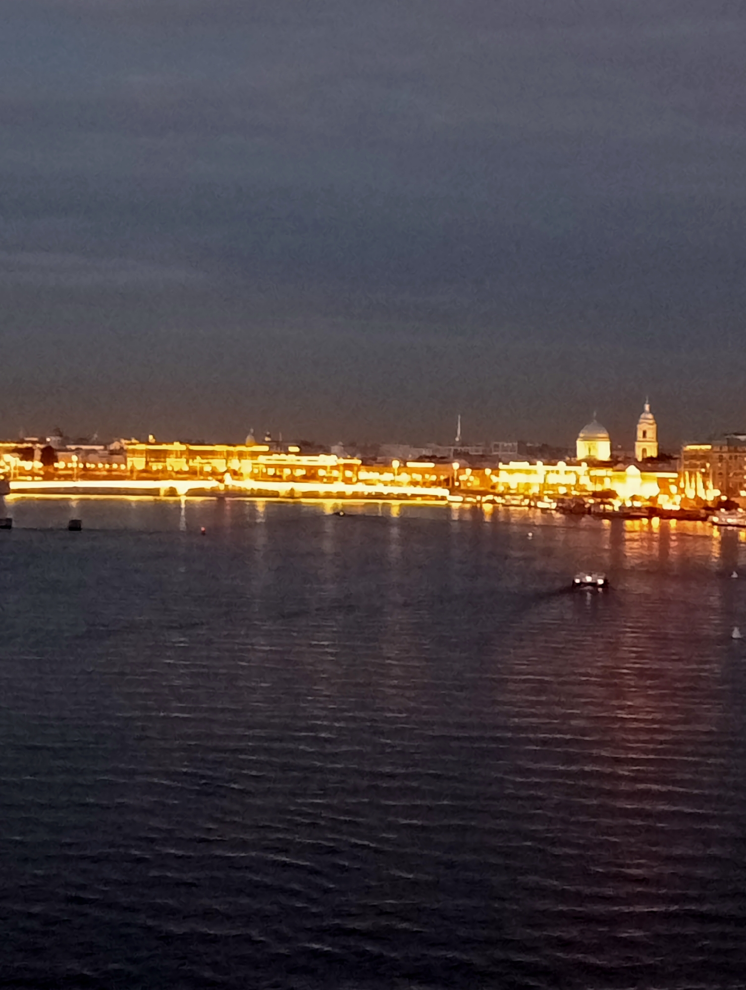 The image depicts a wide shot of the Neva River in Saint Petersburg, Russia, at dusk or night. The city skyline is illuminated by warm, yellow lights, creating reflections on the water's surface. Prominent structures visible include domed buildings, likely churches or governmental institutions, and a long, continuous line of buildings along the embankment. Several boats are present on the river, including a larger vessel with lights in the middle distance and smaller, white buoys or boats scattered further away. The sky is dark and cloudy, with a gradient of dark gray and deep blue. The water is calm, with gentle ripples and the shimmer of the city lights. The overall scene conveys a sense of tranquility and the beauty of the city at night.