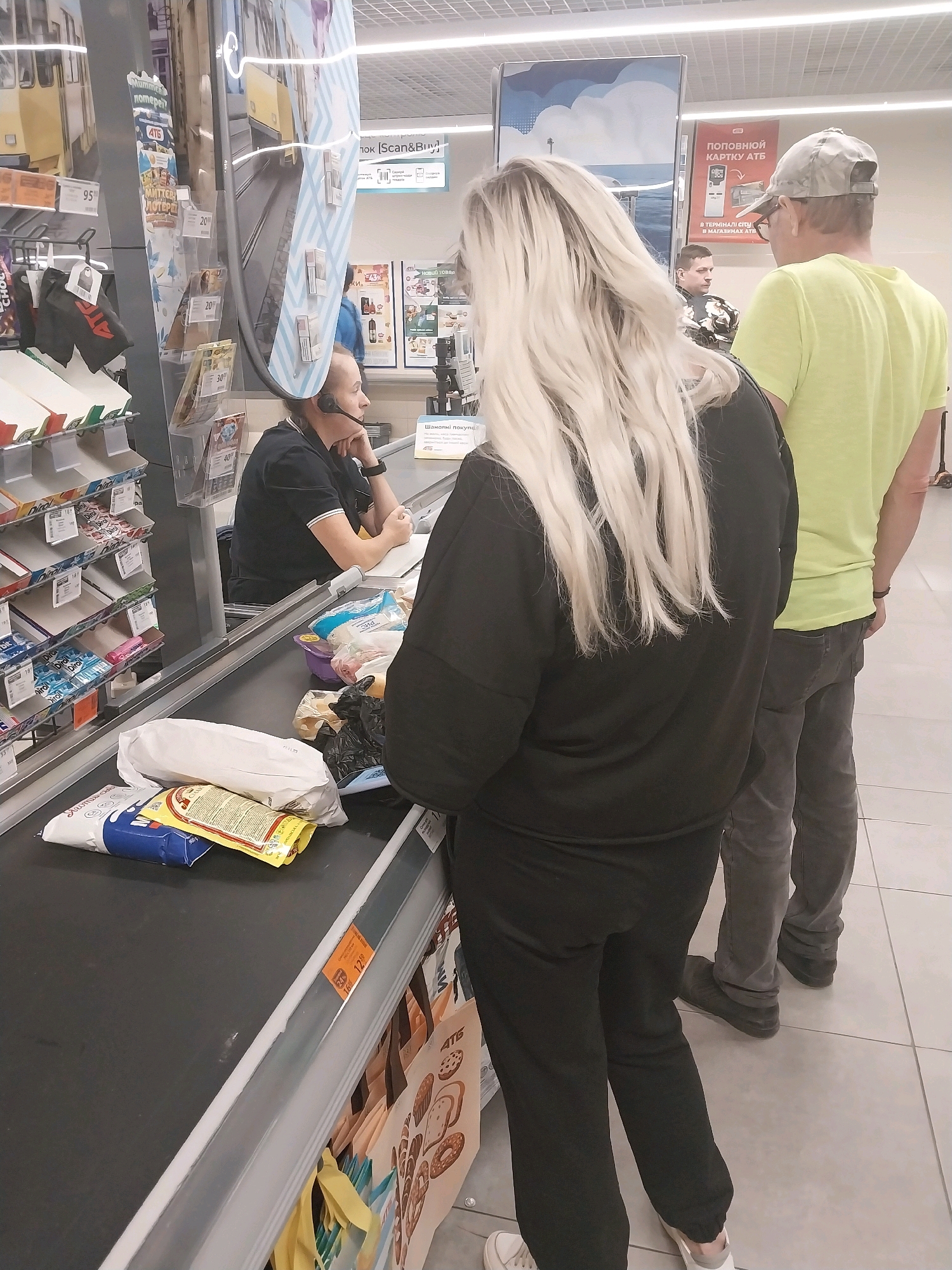 The image depicts a checkout line inside a supermarket in Dnipro, Ukraine. A cashier, wearing a headset, is visible behind a counter. In front of the cashier, on the conveyor belt, are various grocery items including bags, a loaf of bread wrapped in white paper, and other packaged goods. A woman with long blonde hair, wearing a black outfit, stands at the conveyor belt. Behind her, a man in a lime green t-shirt and a cap is waiting in line. The environment suggests a typical grocery store setting with shelving and signage visible in the background.