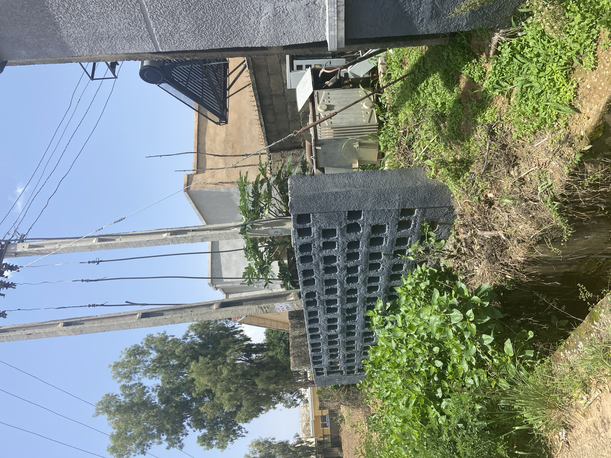 The image depicts an outdoor scene, likely in a residential or semi-industrial area. A prominent feature is a grey, hollow concrete block wall in the foreground, partially covered by lush green vegetation. Behind this wall, an electrical transformer is visible, surrounded by more greenery and dry debris. In the background, there are utility poles with overhead wires, a solar water heater mounted on a building, and several buildings with varying architectural styles and finishes. A large tree with green foliage stands to the left, and the sky is clear and blue. The overall impression is one of an urban environment with visible infrastructure and vegetation. The specific mention of Jos, Nigeria, is not directly supported by visual elements within the image itself.