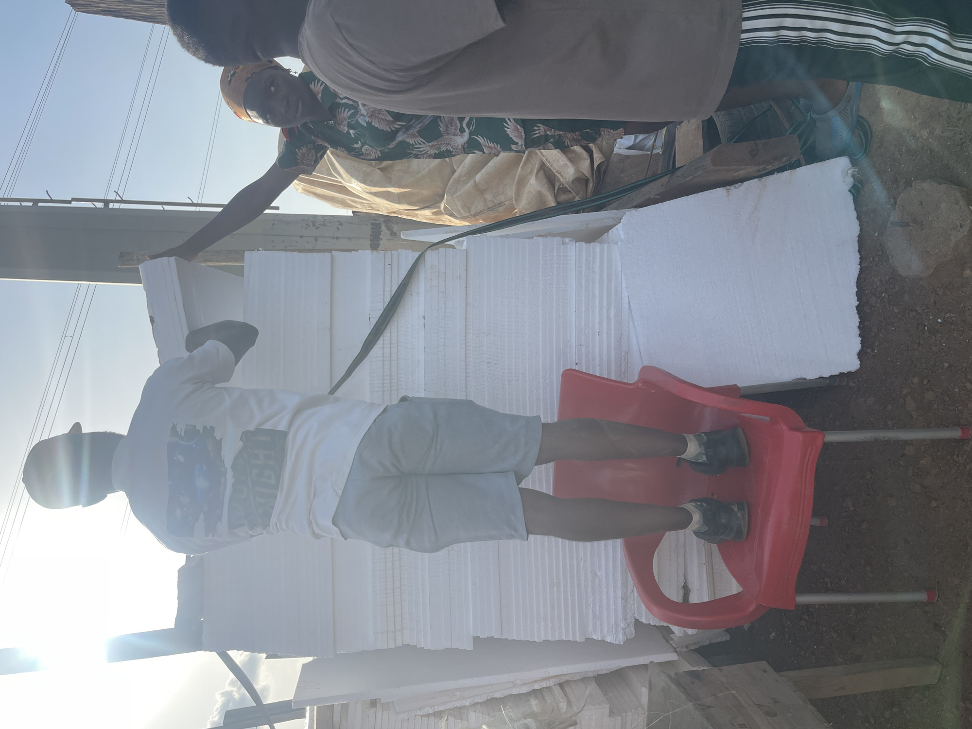 A man in a white T-shirt, gray shorts, and a dark cap stands on a red plastic chair, stacking white styrofoam sheets. He appears to be working with two other individuals, one wearing a floral patterned shirt and the other a tan shirt with green striped pants. The location appears to be an outdoor area in Abuja, Nigeria, given the local environmental details.