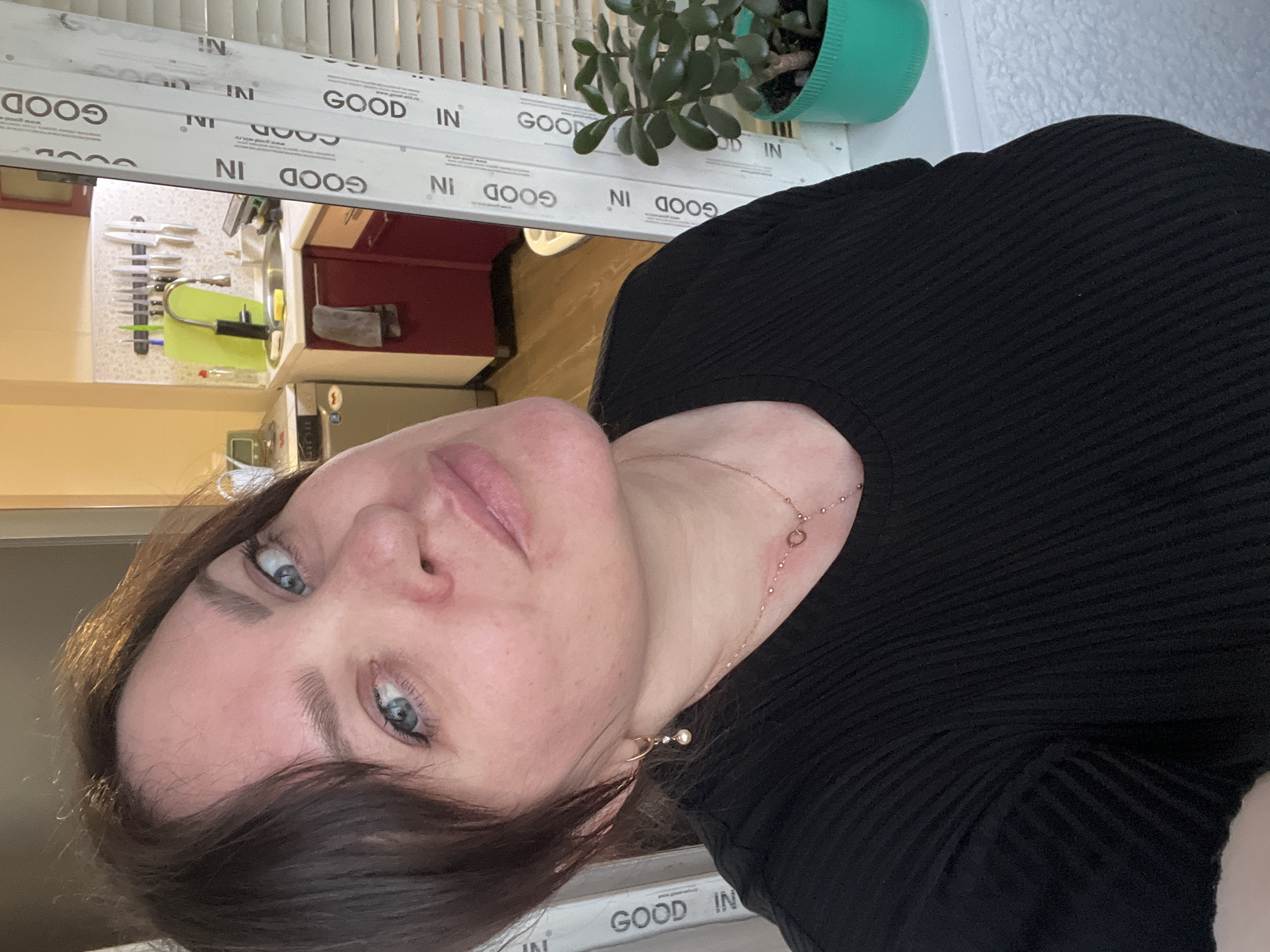 A woman with fair skin, blue eyes, and dark hair is depicted in a close-up selfie. She is wearing a black, ribbed top. A delicate gold necklace adorns her neck, with a circular element visible. The background shows a kitchen setting. A window with white frames and the repeated words "GOOD IN" is visible. On the windowsill, there is a green potted plant with succulent-like leaves. A kitchen sink area is observable, with a cutlery rack and other kitchen utensils visible. Cabinets in dark red are also present. The scene appears to be indoors, presumably in a home kitchen. There is no indication of location in the city of Noviy, Russia.