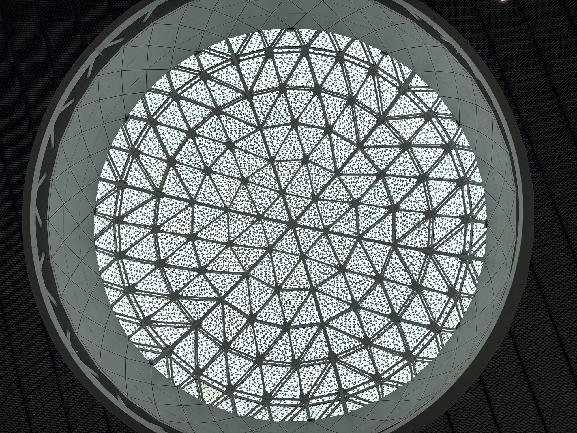 The image presents a striking upward view of a large, circular architectural feature, likely a dome or skylight, within a modern building in Moscow, Russia. The central element is a vast, circular skylight composed of a complex geodesic pattern formed by numerous interconnected triangular panes. Each of these triangular sections is filled with a repeating, intricate pattern of smaller, translucent diamond shapes, which filter and diffuse light from above.
Surrounding this brightly lit, patterned circle is a substantial, lighter grey ring. This ring appears to be a structural or decorative element, possibly a frame or part of the dome's support, and features a smooth, segmented surface. Beyond this light grey ring, the ceiling transitions to a much darker, textured surface, resembling a dark grey or black metal mesh or perforated panels. This dark surrounding contrasts sharply with the luminous central dome.
No people or identifiable activities are visible in the static architectural shot. The bright light streaming through the dome suggests it is daytime. The overall aesthetic is one of modern design, emphasizing geometric patterns, light, and contrasting textures and tones. There is no visible text.