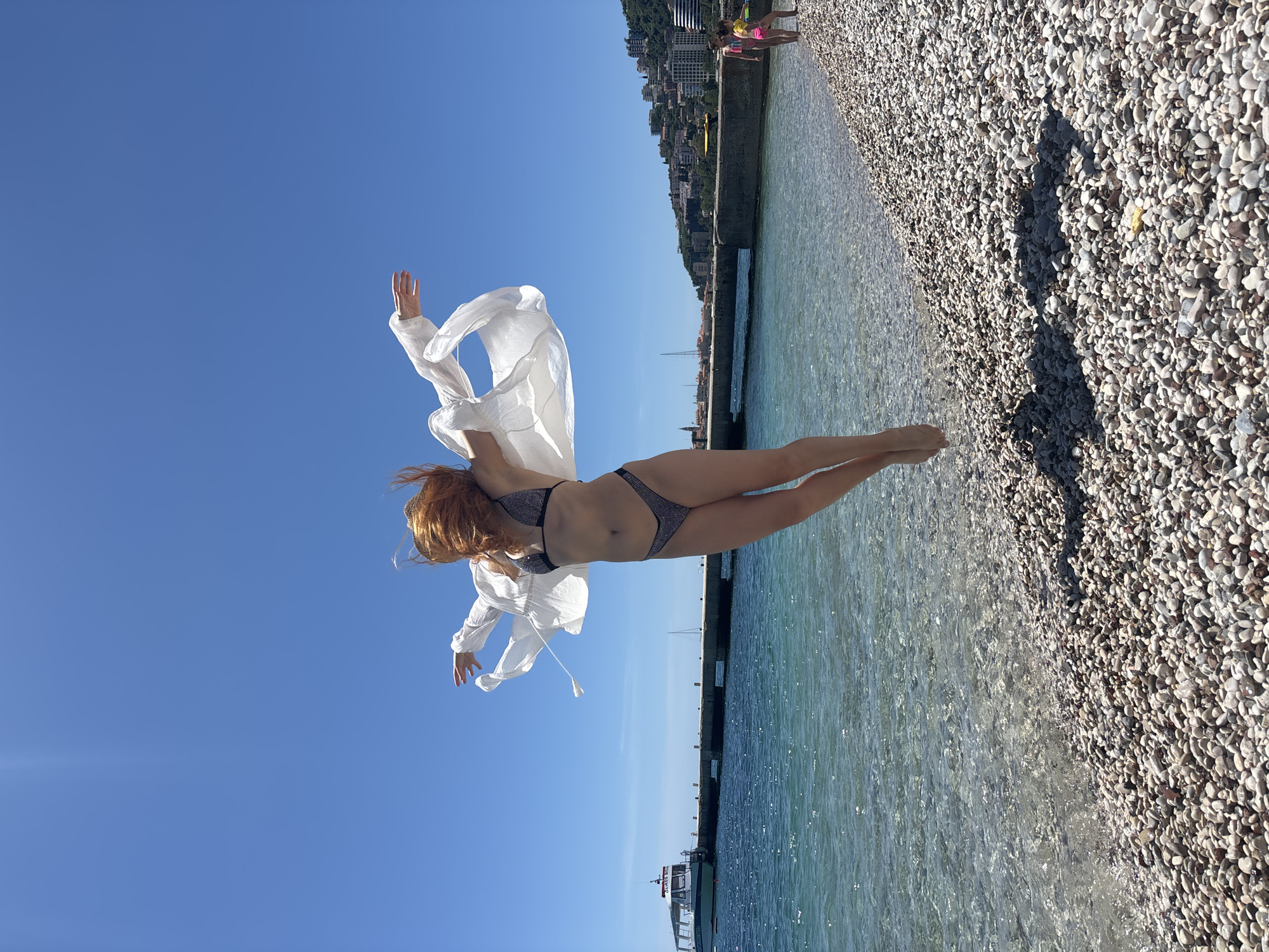 A woman with long red hair is suspended in the air above the water, wearing a bikini and a white shirt that is open and flowing. Her arms are raised. The sky is blue, and the water is clear. There is a stone beach and a concrete pier/wall to the right and a coastline with buildings in the distance. A small boat is visible on the water near the pier. Two people are standing on the beach in the distance. The location appears to be a beach area in Budva, Montenegro, based on the architecture and coastal features.