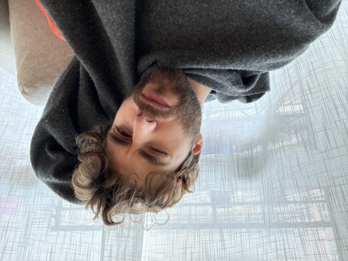 The image displays a close-up, head-and-shoulders view of a single male individual in an indoor setting within Paris, France, likely an apartment or room. The man has medium brown, curly hair and a dark brown, well-groomed beard and mustache. He is looking directly at the camera with his right eye open and his left eye predominantly closed, creating a winking or squinting expression. His eyebrows are slightly furrowed. He is wearing a dark grey, textured garment, possibly a hooded sweatshirt or a blanket, draped over his left shoulder and partially obscuring his body. A portion of a light beige or tan sofa or armchair is visible behind his right side, with a faint orange detail between the grey garment and the furniture. The background features a window covered by a translucent white curtain with a distinctive grid-like woven pattern. Through the curtain, indistinct architectural forms suggesting urban buildings are discernible, indicating a view of the cityscape.