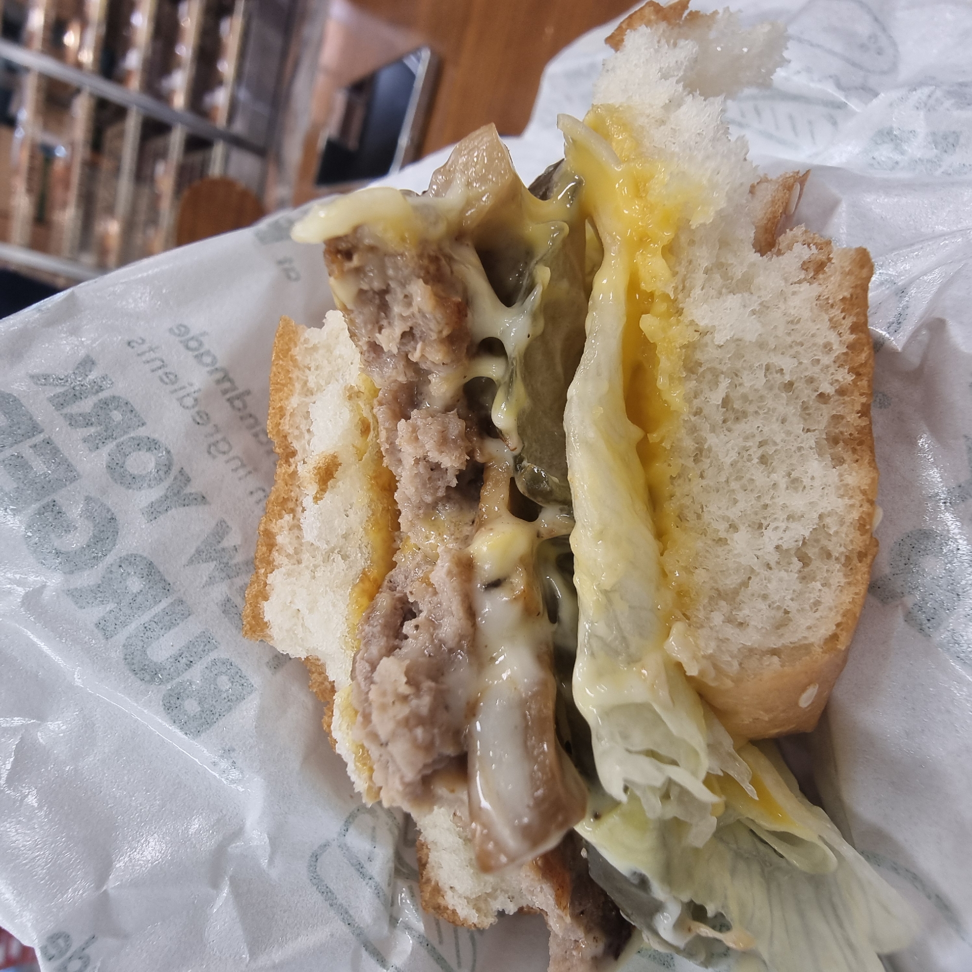 A close-up, eye-level shot captures a partially eaten burger, wrapped in white paper with embossed text that is partially visible. The burger, with its top bun removed, reveals layers of meat, melted cheese, lettuce, and pickles. The texture of the burger's ingredients, including the crumbled meat patty and gooey cheese, is evident. The background is softly blurred, showing shelves stocked with food items, suggesting a retail or food service environment. The lighting is bright, highlighting the food. The exact location within Wonju-si, South Korea, is not discernible from the image itself.