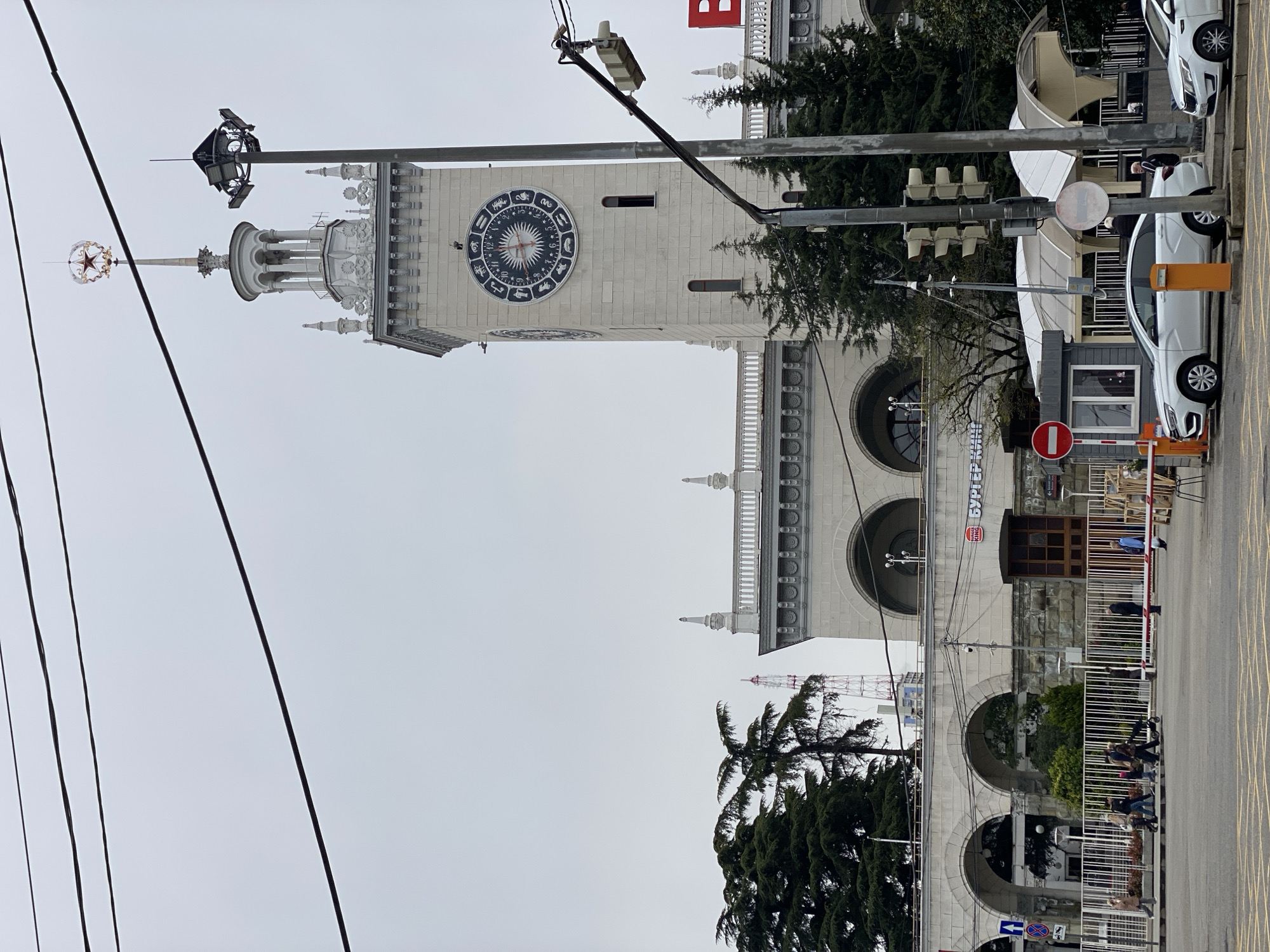 The image shows a view of the Sochi Sea Port building in Sochi, Russia, on an overcast day. The focus is on the clock tower of the building, which features a large circular clock with astrological symbols. Above the clock tower is a spire topped with a star. There are utility poles and wires in the foreground. The main building section features arched windows and decorative elements. There are a few people seen walking on the sidewalk near a gated area with barriers. Cars are parked along the side of the road adjacent to the building. Evergreen trees are seen in front of the building.