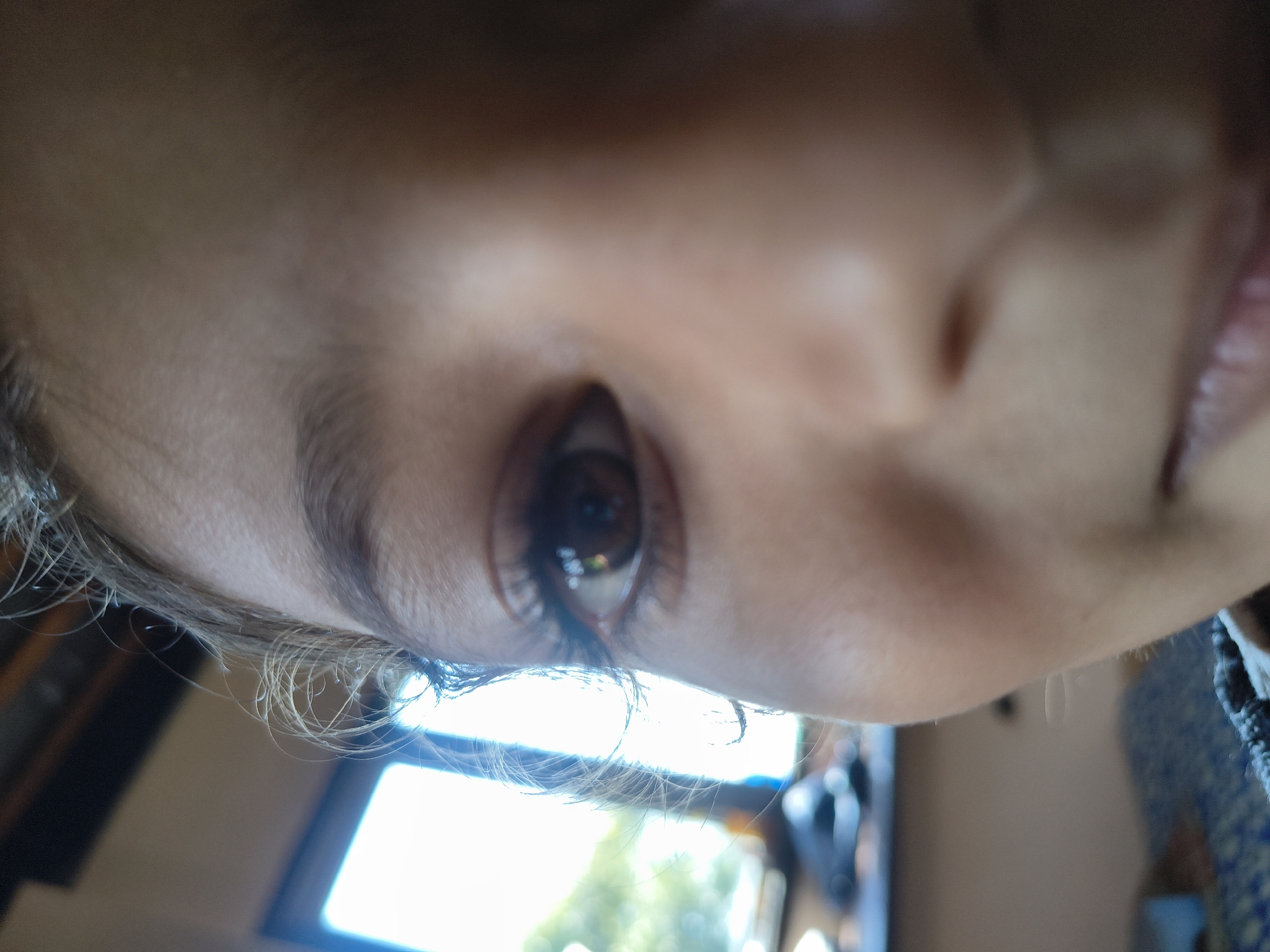 A close-up, low-angle, eye-level shot captures the right side of a child's face, focusing on their eye and cheek. The child has brown eyes, long eyelashes, and a light complexion. Their hair, light brown and wispy, frames their face. The background is blurred but suggests an indoor setting with a window and foliage visible outside. The image quality is grainy, and the focus is soft, creating an impressionistic and intimate portrait. The child's mouth is slightly open, revealing the lower lip. The context of Kandi, India, is not evident from the visual elements of the image.