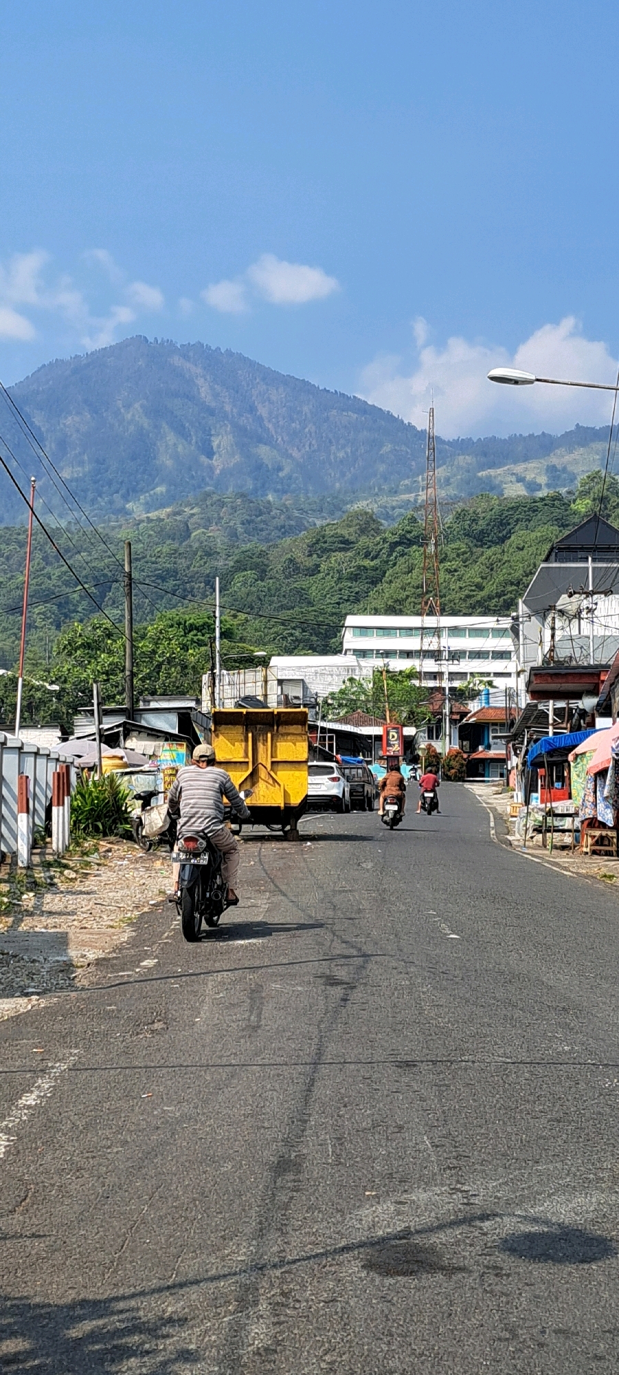 A vertical, eye-level, medium shot shows a paved road in a village setting with a mountain range in the background. A man wearing a striped shirt and a cap rides a motorcycle away from the camera, wearing tan pants and black shoes. A yellow dumpster truck is parked to his left. Beyond the truck are several buildings, some of which appear to be shops. A tall telecommunications tower is visible in the middle ground, with a streetlamp post on the right side of the image. Two other motorcycles are further down the road, one with two riders and another with a single rider. The sky is clear and blue with a few scattered clouds. The road is asphalt with some wear and tear, and a white line is visible on the left side. The surroundings are hilly and covered in dense green vegetation. This scene is likely located in Prigen, Indonesia, given the tropical climate and mountainous terrain.