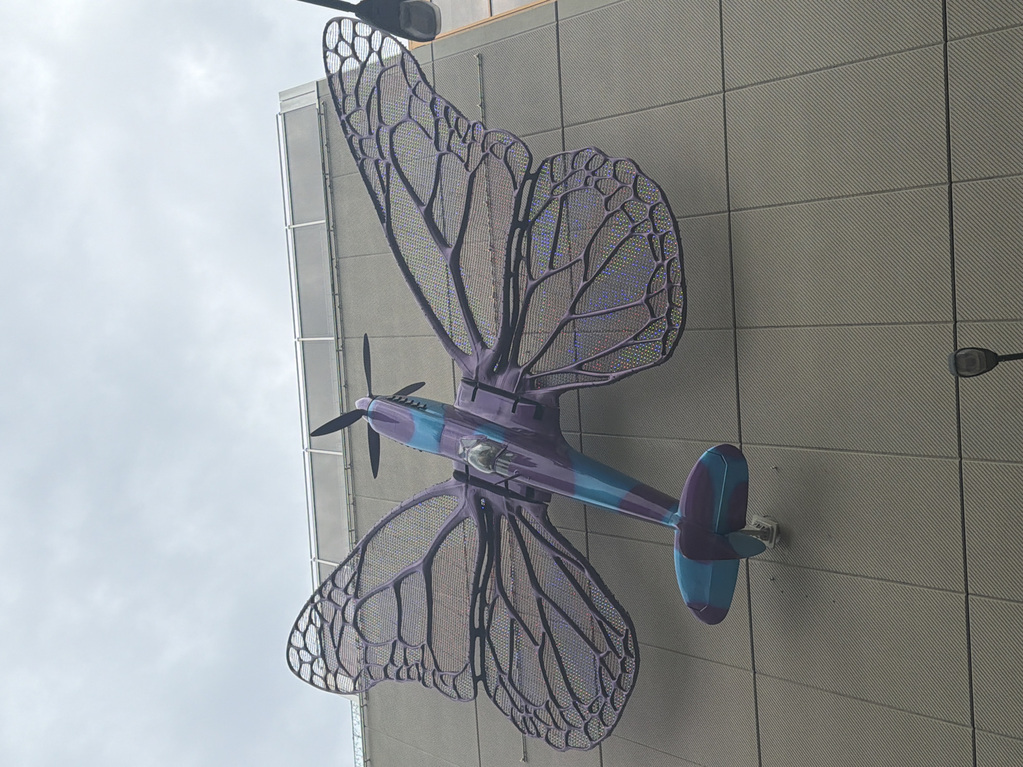 An outdoor sculpture of an airplane with butterfly wings is mounted on the side of a modern building. The sculpture is predominantly purple and teal. The airplane's fuselage is painted with purple and teal stripes, and it has a propeller at the front. The wings are large and resemble butterfly wings, with a black, veined structure and a shimmering, iridescent mesh filling the spaces between the veins. The building's facade is a light gray, textured material, with horizontal lines formed by the panels. Above the sculpture, a gray, cloudy sky is visible. A streetlamp is visible in the lower right corner. The sculpture is located in Prague, Czechia, at the Florentinum office building complex.