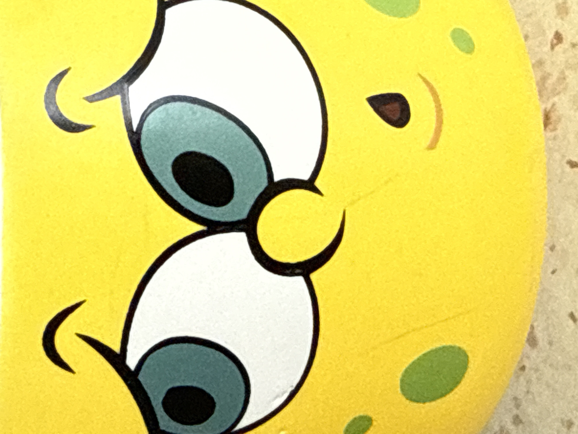 The image is a close-up, eye-level, full-shot of a yellow, cartoon character's face, likely Spongebob Squarepants, against a textured, light-colored surface. The character has large, white eyes with teal irises and black pupils, angled upwards and to the left, giving it a slightly concerned or inquisitive expression. Black, curved eyebrows are positioned above each eye. The character's mouth is a small, smiling crescent shape, and above it is a small, dark brown, teardrop-shaped nose. The yellow surface of the character's face is dotted with irregular, green ovals, suggesting a textured or porous appearance. The lighting appears to be even, with no harsh shadows. The background is out of focus, but the textured surface suggests it could be a table or countertop. The image does not provide any information about the location in Tbilisi, Georgia, or any other specific location.