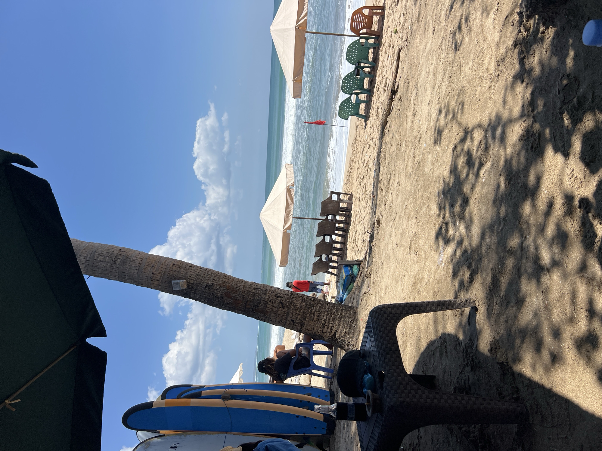 This image depicts a beach scene in Kuta, Indonesia, under a clear blue sky with scattered clouds. In the foreground, a dark, woven plastic table sits on the sand, with a dark bottle and a black cap on it. A large, dark green umbrella partially shades the area. To the left, several surfboards, predominantly blue and yellow, are propped up. A person sits on a blue plastic chair facing the ocean, and another person in a red shirt stands near the water's edge. The middle ground features two tan beach umbrellas providing shade for rows of plastic chairs. A small red flag is planted in the sand near the water. The ocean is visible with moderate waves breaking on the shore. The sand appears light brown and there are shadows from trees or other overhead structures cast on the ground.