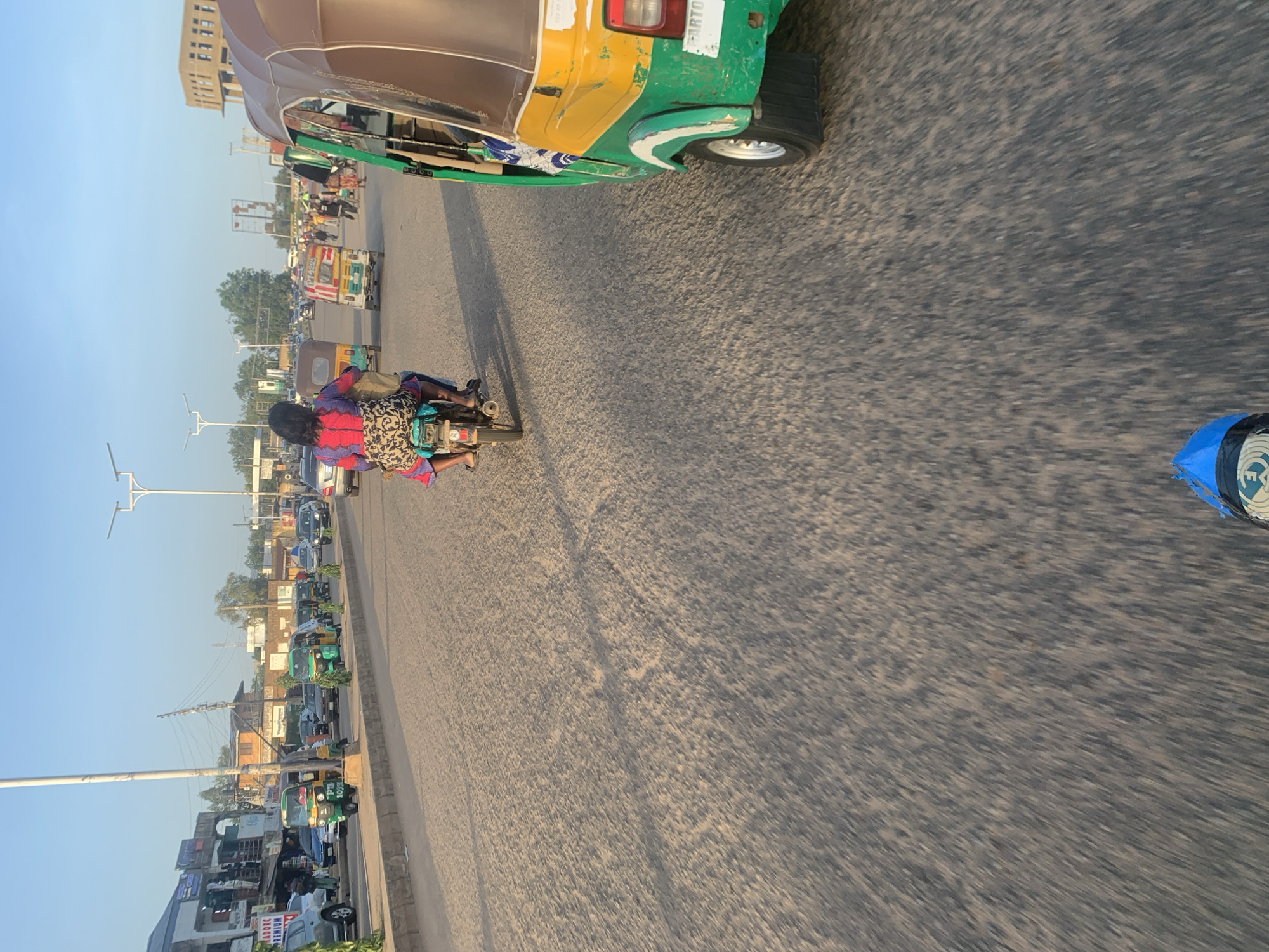 The image depicts a street scene in Kabong, Nigeria, characterized by moderate vehicular traffic and pedestrian activity. In the foreground, a green and yellow auto-rickshaw is visible, positioned on the right side of the road. Directly ahead, a motorcycle carrying two individuals is moving away from the viewer. The motorcycle rider is wearing a blue and red patterned top and a dark skirt, while the passenger is partially visible behind them.
The road surface is asphalt, showing signs of wear and tear. Along the left side of the road, a row of buildings with shops and storefronts is present. Several other auto-rickshaws, cars, and motorcycles are scattered throughout the scene, indicating ongoing movement. The sky is clear and blue, suggesting fair weather. The overall impression is of a busy urban thoroughfare.
