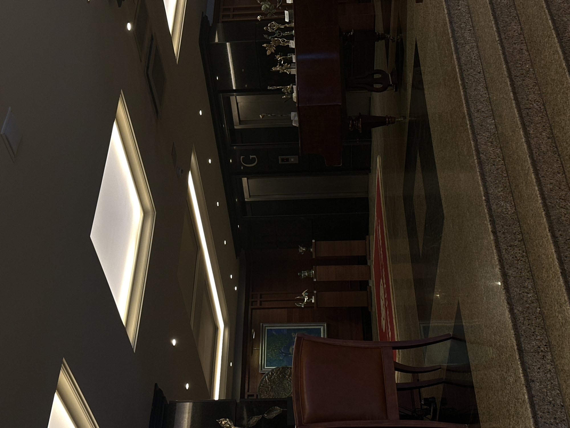 The image captures the interior of what appears to be an elegant and dimly lit lobby or hallway within a building, likely in Sofia, Bulgaria. The perspective is slightly elevated, looking down into the space.
On the left, a striking architectural wall features geometric patterns defined by linear LED strip lighting embedded in coves, casting a warm glow. Numerous small, circular recessed spotlights are also visible on the wall and ceiling, providing additional illumination.
In the mid-ground, a dark wooden grand piano stands prominently, adorned with several golden, intricate decorative objects or trophies. Behind the piano, a dark-paneled wall reveals an elevator entrance. A large, illuminated white letter "G" (likely indicating "Ground floor") is visible on the elevator door, along with a call button. Further back, more dark wood paneling is seen, possibly with shelves displaying small items, and a framed painting or artwork.
To the right, a grand staircase with wide, speckled granite or marble steps ascends diagonally. A red carpet runner is partially visible on the floor leading towards the staircase. In the foreground, a dark wooden chair with a brown upholstered seat and back is positioned, facing away from the viewer. Another framed picture or artwork hangs on the wall near the chair.
The overall atmosphere is quiet and sophisticated, with no people present. The primary source of light comes from the architectural lighting fixtures, suggesting an evening setting or an intentionally mood-lit interior. The rich materials like polished stone, dark wood, and warm lighting create an opulent yet serene environment.