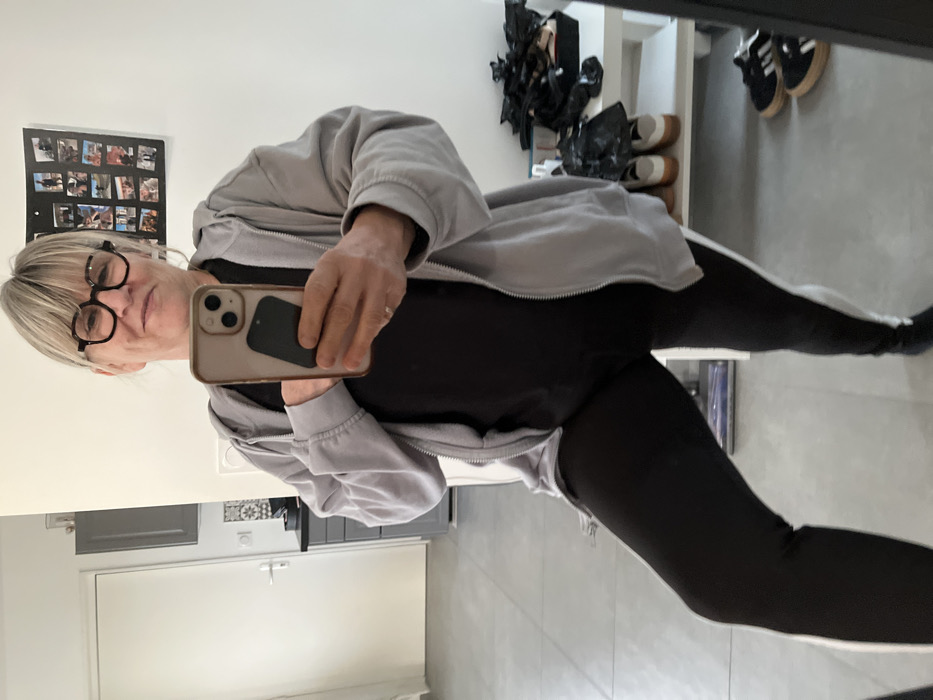 The image is a mirror selfie showing a person, likely female, from the mid-thigh up, taken indoors. The perspective is rotated 90 degrees to the left.
The person has blonde hair, parted on their left side (right in the image), and is wearing black-framed, round glasses. They have a light complexion and a faint, closed-mouth smile. Their right hand (appearing on the left in the rotated image) holds a light-colored smartphone with a darker, rectangular object (possibly a card holder or another phone) attached to its back. A simple, thin ring is visible on the ring finger of the hand holding the phone.
The person is dressed in a casual outfit consisting of a light grey zipped hoodie, worn open, revealing a black top underneath. They are also wearing black leggings with a distinct white stripe running down the outer side of each leg.
The background, which is a reflection in the mirror, shows different areas of an indoor space. To the person's left (upper-left in the rotated image), there is a black framed display or photo collage on a light-colored wall, containing several small photographs arranged in a grid. To the person's right (upper-right in the rotated image), there is a shelving unit with several pairs of shoes. Some shoes are dark with white soles and stripes (resembling Adidas Gazelles), while others have light-colored soles and darker uppers. Also on the shelf are some crumpled, dark objects, possibly bags.
Below the person's reflection (lower part of the image), there is a reflection of what appears to be a different room or area. This area includes light grey or white cabinets, a light-colored countertop, and a backsplash with a dark and light patterned tile design. The floor is a light, neutral color, possibly light grey tile or concrete. The overall setting suggests a home environment. No discernible text is visible in the image.