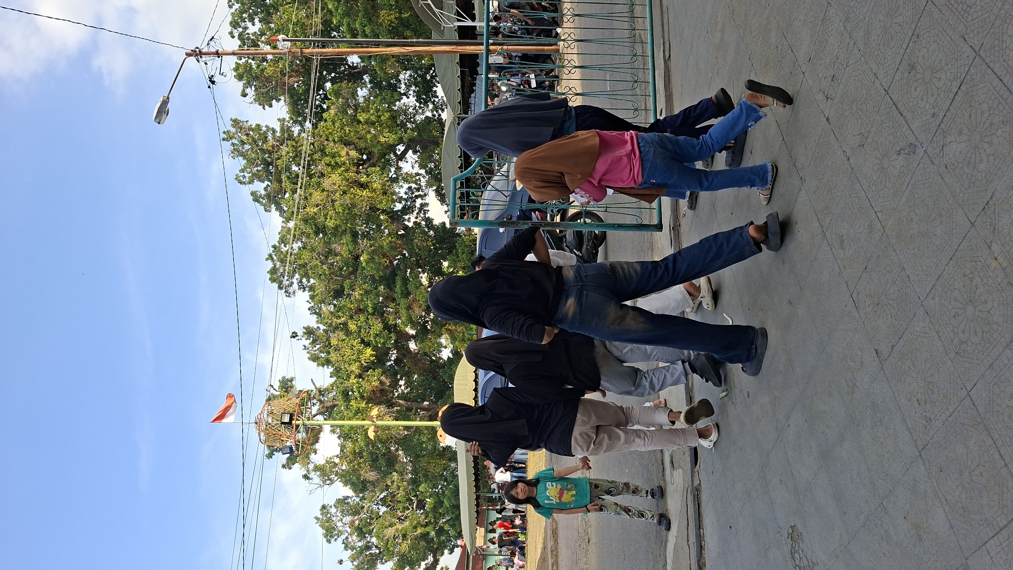 This is an outdoor, eye-level, medium shot of several people walking on a paved surface in Lhokseumawe, Indonesia. The group includes at least six individuals, a mix of adults and children, some wearing hijabs. In the background, there are trees, a street light, power lines, a fence, and some buildings with awnings. A red and white flag, likely the Indonesian flag, is visible on a pole behind a circular structure. The lighting suggests it is daytime, possibly late afternoon, with shadows indicating the sun is lower in the sky. The ground is a patterned pavement, and the overall atmosphere appears casual, with people moving in the same direction.