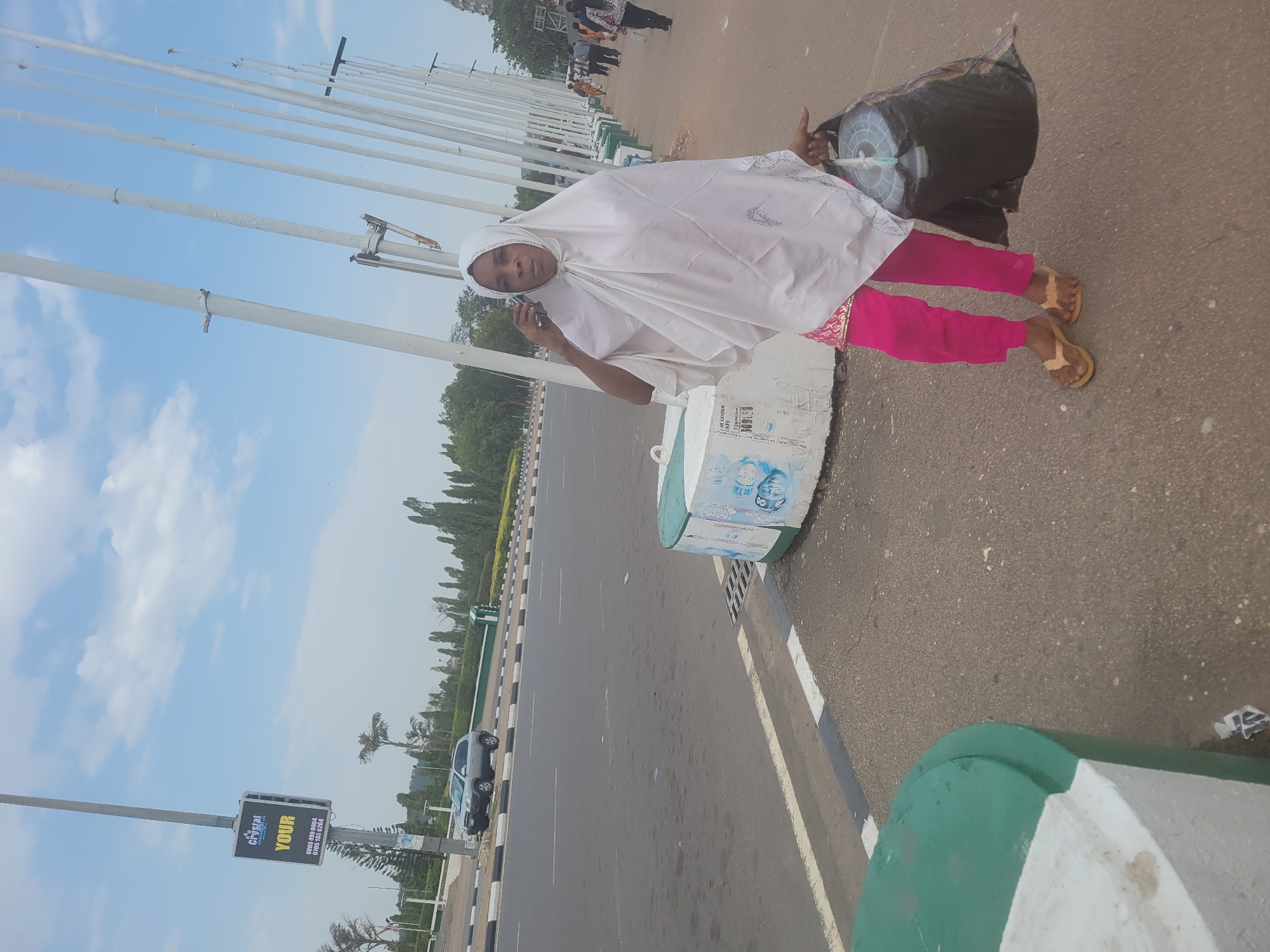 The image shows a woman standing on the side of a road. She is wearing a white headscarf, a white top, and pink trousers. She is holding a cell phone to her ear with her left hand and she is holding a large black bag over her right shoulder. The road is wide and has several lanes. There are poles on the left side of the road. In the background, there are trees and buildings. There is a billboard on the left side of the road for "The Crystal". Based on the visual clues, the location appears to be in Abuja, Nigeria, judging by the architecture and environmental features.