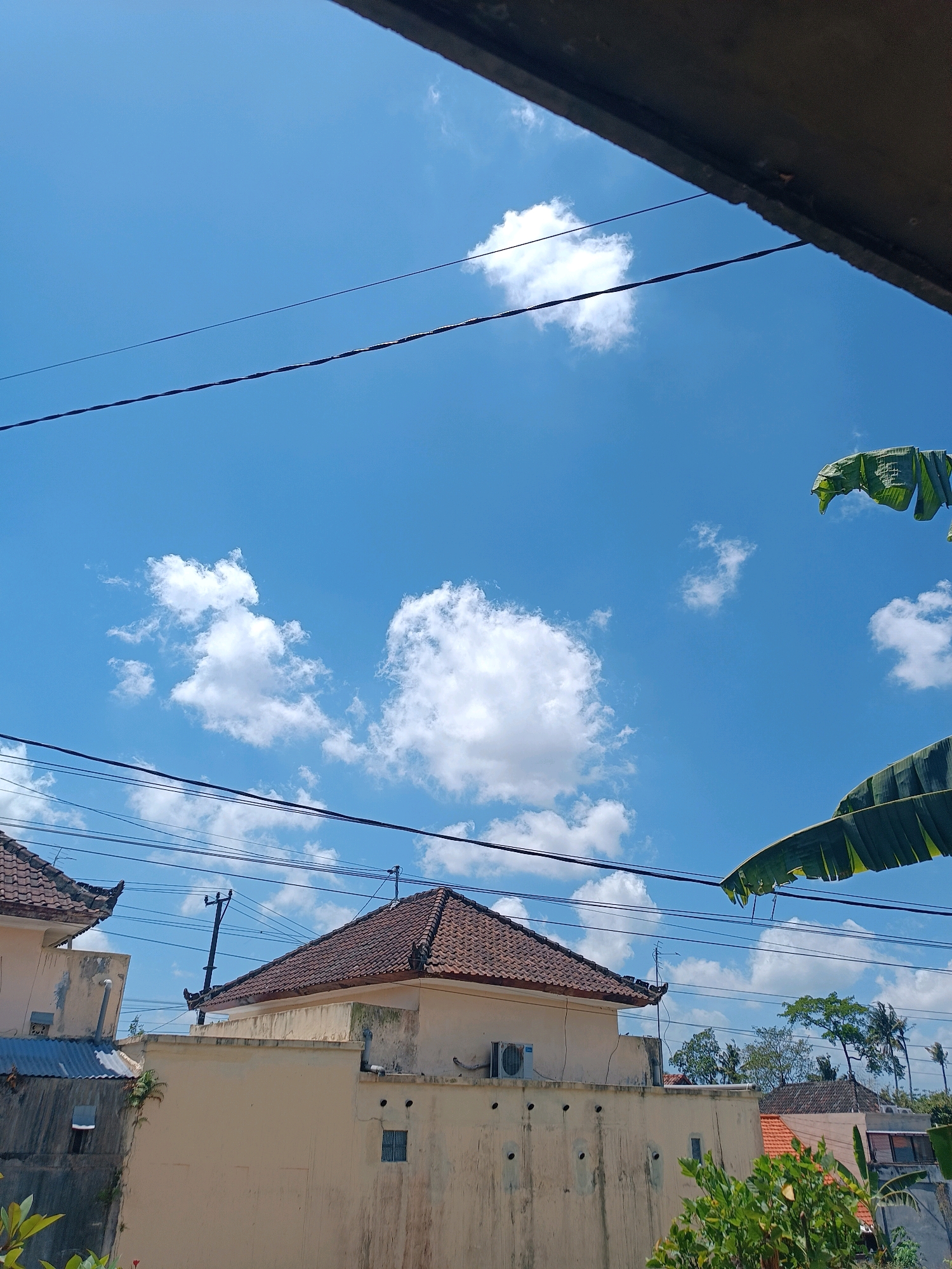 The image captures a bright, sunny day in Tabanan, Indonesia, with a clear blue sky dotted with fluffy white clouds. The foreground is partially obscured by a dark, angled structure, possibly a roof overhang, and several power lines stretch across the frame. Below the sky, the scene transitions to a residential area. Two houses with traditional tiled roofs are visible, one with a lighter colored exterior and another to its left with a darker, corrugated metal roof. An air conditioning unit is mounted on the wall of the lighter colored house, and ventilation holes are visible. In the distance, lush green trees and more rooftops suggest a densely populated area. Large, green banana leaves frame the top right and bottom right of the image, adding a tropical element to the scene. There are no people visible in the image. The overall impression is a glimpse into the everyday life of a town in Indonesia under a clear sky.