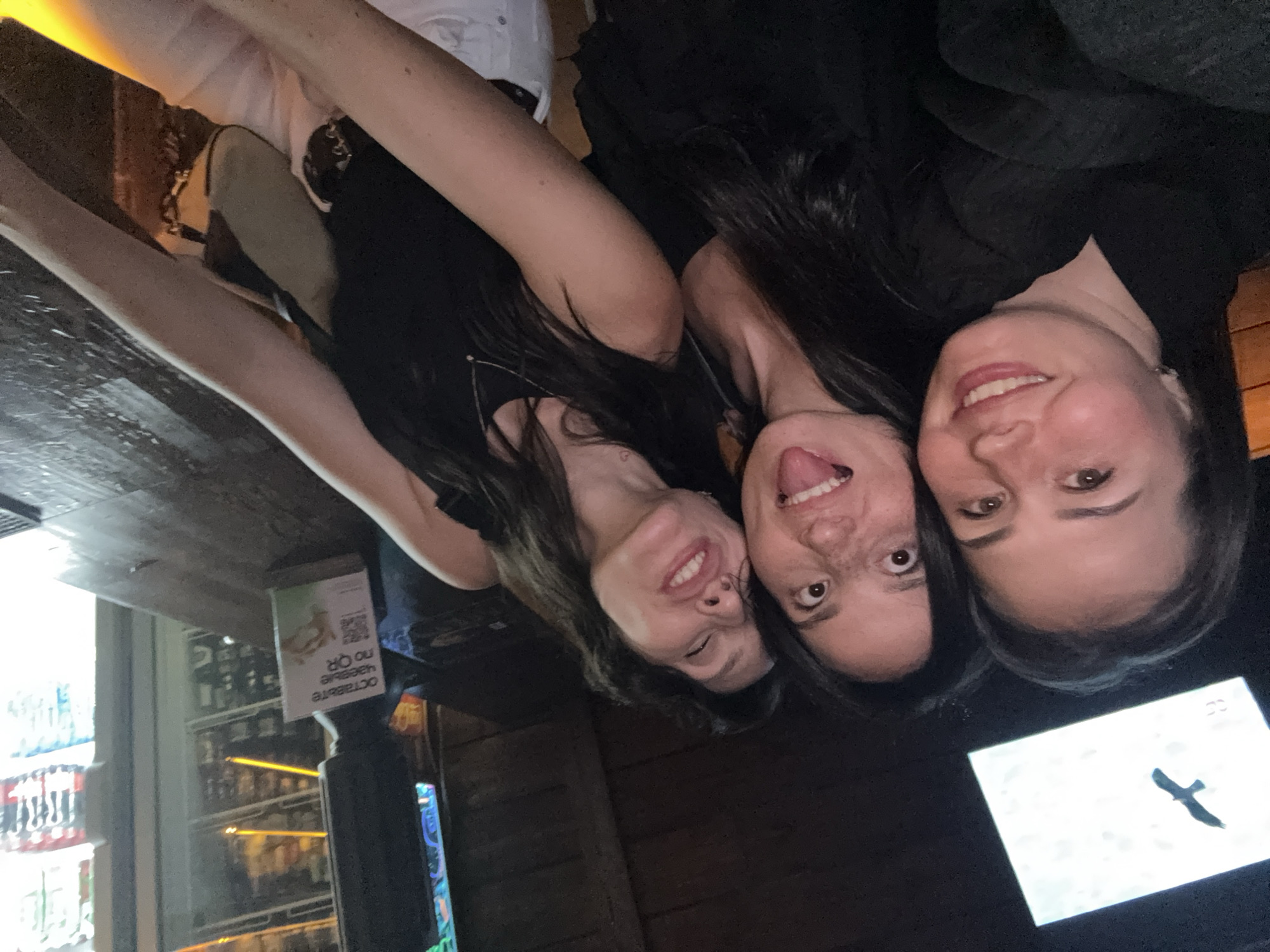 Three women are posing for a selfie inside what appears to be a bar or restaurant. The woman on the left is smiling and winking. The woman in the center is sticking her tongue out. The woman on the right is smiling directly at the camera. All three women have dark hair and are wearing dark-colored tops. In the background, there is a display case with beverages, a promotional sign, and a television screen. The television screen appears to have a black bird on a white background. The text on the sign is upside down, and partly illegible, but the word "OCTOBER" can be made out. Based on the language used on the sign, the location of the photograph is most likely in Russia, more specifically, Chaikovsky.