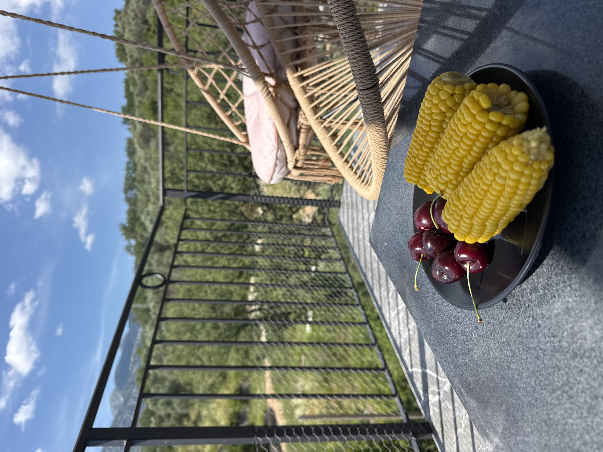 On a balcony overlooking a verdant hillside in Sustaš, Montenegro, a black plate holds three ears of corn and several ripe cherries. The corn, a vibrant yellow, is arranged diagonally across the plate, with the kernels clearly visible. The cherries, a deep red, are clustered to the left of the corn, their stems attached. In the background, a black metal railing separates the balcony from the natural landscape. Beyond the railing, a lush green hillside is dotted with trees, suggesting a Mediterranean climate. In the distance, hazy mountains rise under a bright blue sky with scattered white clouds. Two macrame hanging chairs, one with a pink cushion, are positioned on the balcony, hinting at a place of relaxation and leisure. The overall scene evokes a sense of peaceful outdoor dining amidst a scenic natural setting.