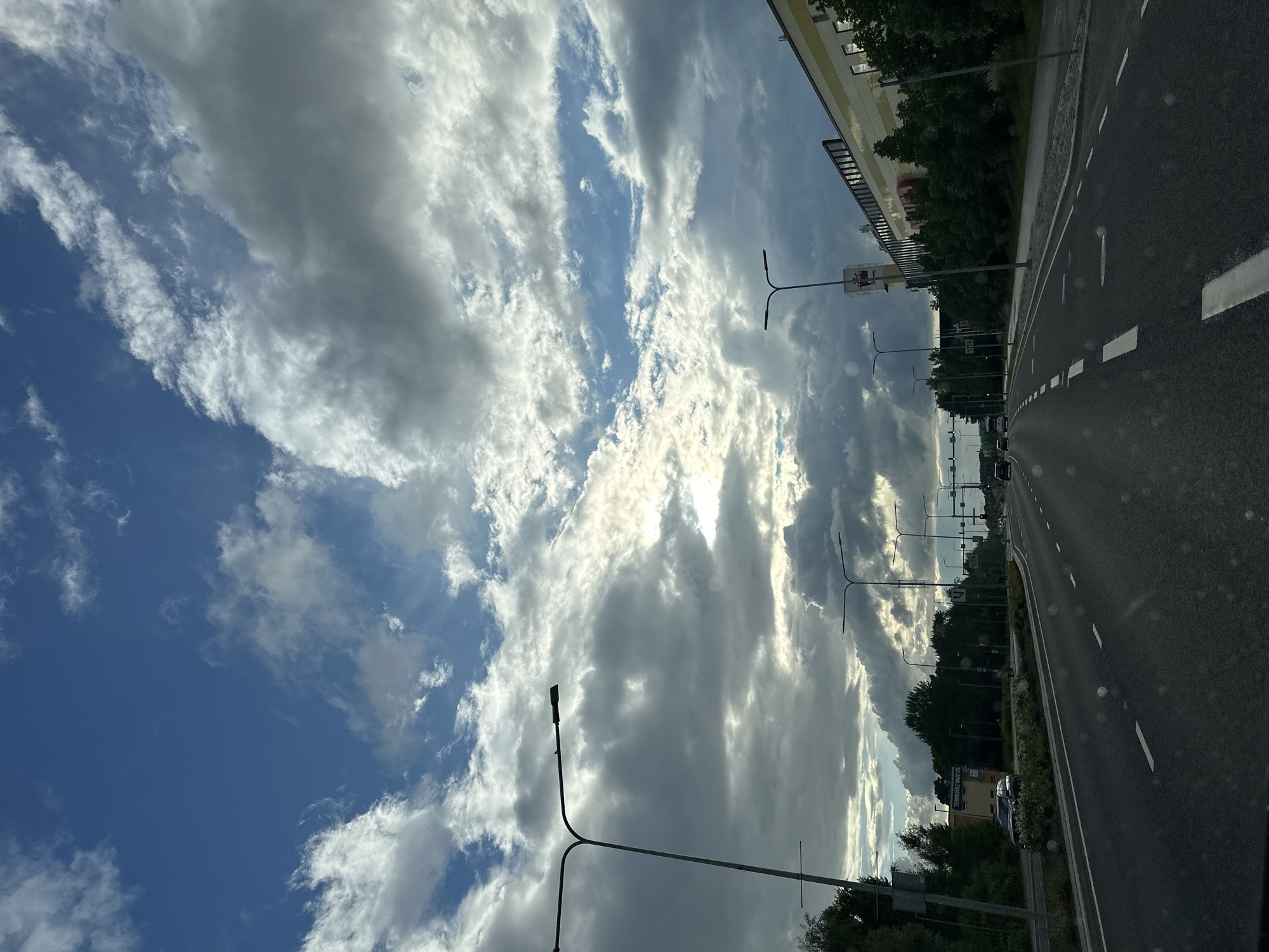 The image depicts a multi-lane road in Tallinn, Estonia, seen from the perspective of a vehicle in motion. The sky is a dramatic mix of blue and dark, heavy clouds, with patches of sunlight breaking through. Several streetlights line the sides of the road, and there are trees and buildings visible in the background. Cars are driving on the road, indicating ongoing traffic. A large, modern-looking building with a yellow facade and dark windows is prominent on the right side of the image. The road itself is asphalt with clear white lane markings. The overall impression is of a slightly overcast, late afternoon or early evening scene in an urban or suburban setting.