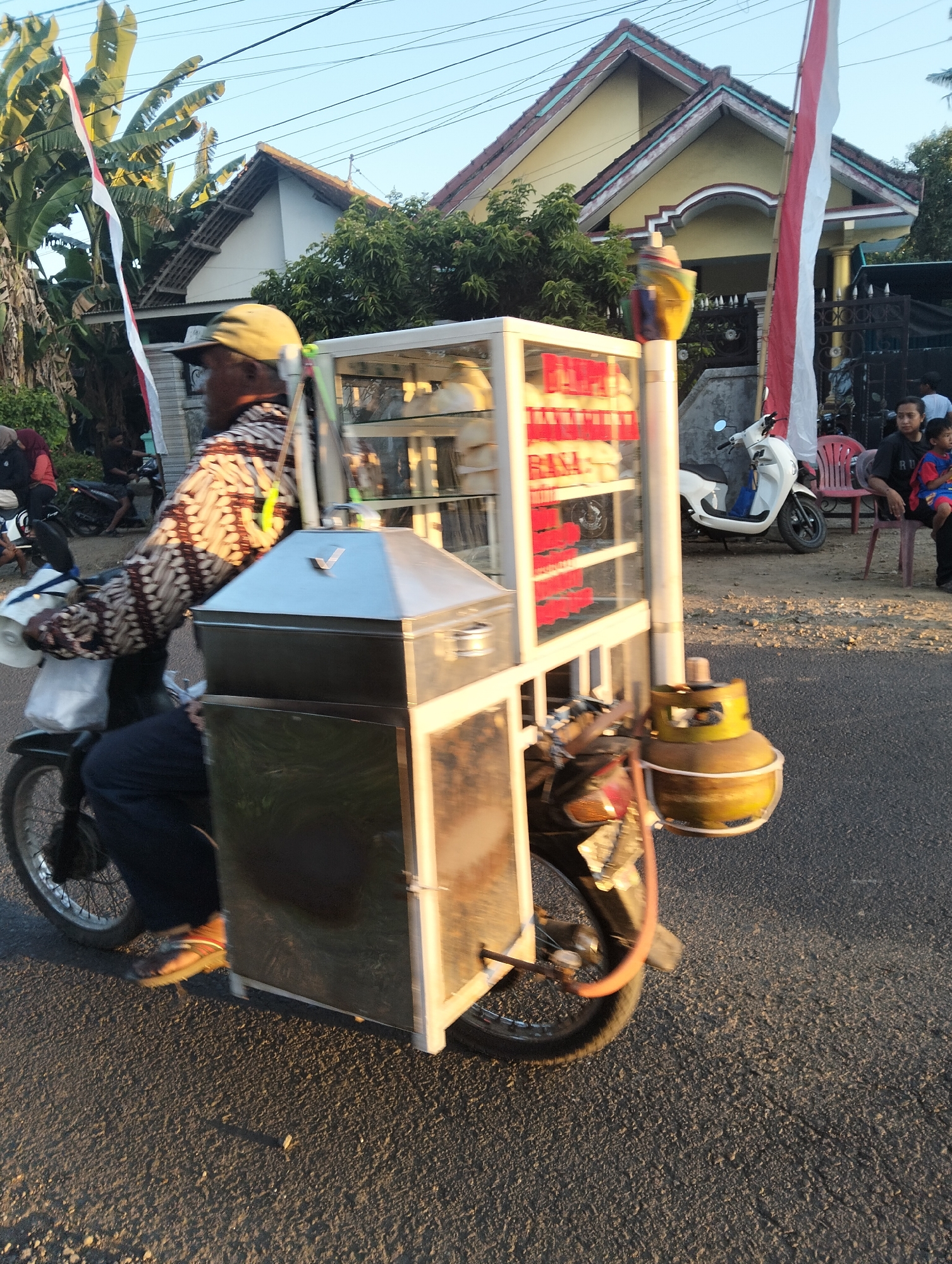 The image depicts a street scene in Sumbersewu, Indonesia, with a street vendor on a motorcycle-pulled cart. The vendor, wearing a cap and a patterned shirt, is riding the motorcycle. The cart is equipped with a glass display case showcasing food items and a propane tank. In the background, a house with a yellow facade and a red-tiled roof is visible, adorned with Indonesian flags. Several people are present, including a woman and child sitting on chairs outside the house, and other individuals near a parked white scooter. The environment appears to be a residential street with trees and overhead wires. The lighting suggests it is late afternoon or early morning.