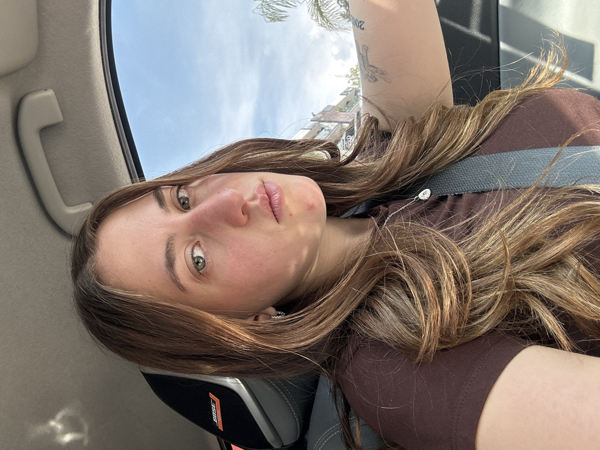 A young woman with light brown hair is in the front seat of a car. She is wearing a brown t-shirt and a seatbelt. She has a tattoo on her upper arm. The interior of the car is visible, including the dashboard, headrest and grab handle. Through the car window, a partial view of buildings and a palm tree suggests an urban environment. While the image provides visual details, it does not offer sufficient evidence to definitively determine the location as Huixquilucan de Degollado, Mexico.