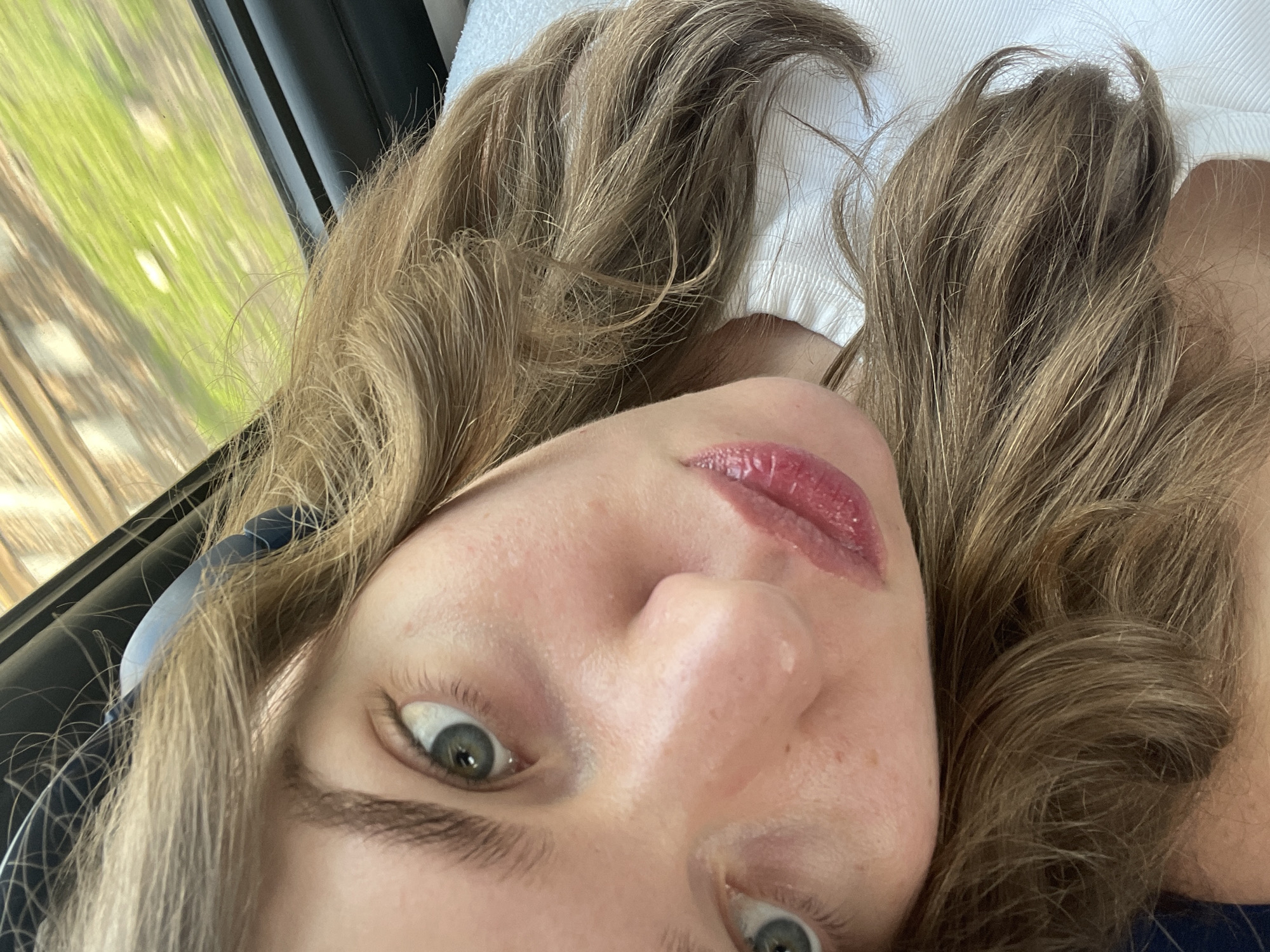A selfie shot, taken from a low angle, shows a woman with light brown, wavy hair and green eyes. She is wearing lip gloss. In the background, visible through a window, are trees and a blurred landscape. The location is not identifiable as Moscow, Russia. The woman appears to be resting her head on a white pillow.