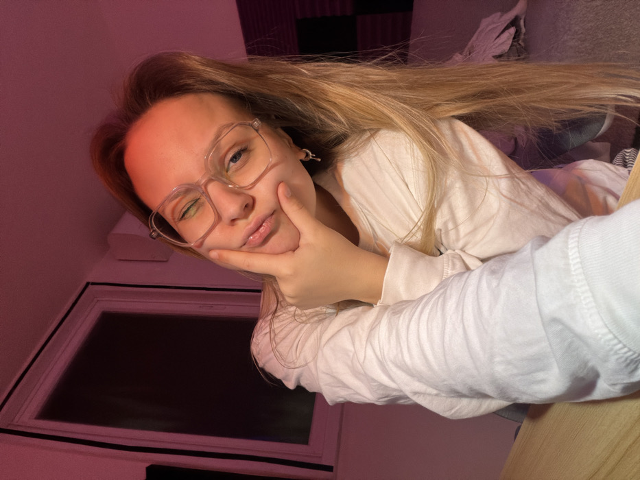 A young woman with long blonde hair and glasses poses with a hand on her cheek, winking at the camera. She wears a white long-sleeved shirt. The background includes a wall with soundproofing panels, a window, and what appears to be bedding. The lighting has a strong pink tint, casting a reddish hue on the scene. The perspective is slightly tilted, giving the image a casual, selfie-like feel.