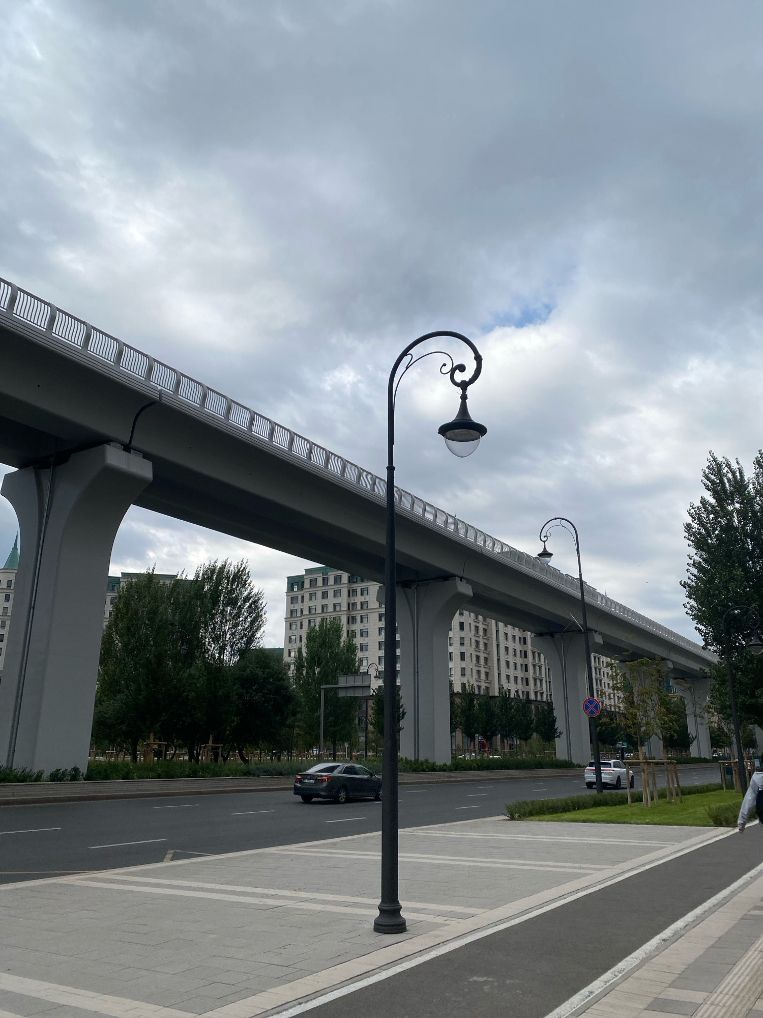 This image captures a street scene in Astana, Kazakhstan, featuring a modern elevated road or bridge structure spanning across the frame. The structure is supported by large concrete pillars, and its railing is visible along the top. Beneath the elevated road, there is a street with traffic, including a dark-colored sedan. Tall, multi-story buildings with distinctive green roofs are visible in the background, suggesting an urban environment. Several trees line the street, and there are pedestrian walkways with decorative paving. Two ornate street lamps are positioned in the foreground, one prominently in the center and another further down the street. The sky is overcast with heavy clouds. A person is partially visible on the right side of the image, walking away from the camera.