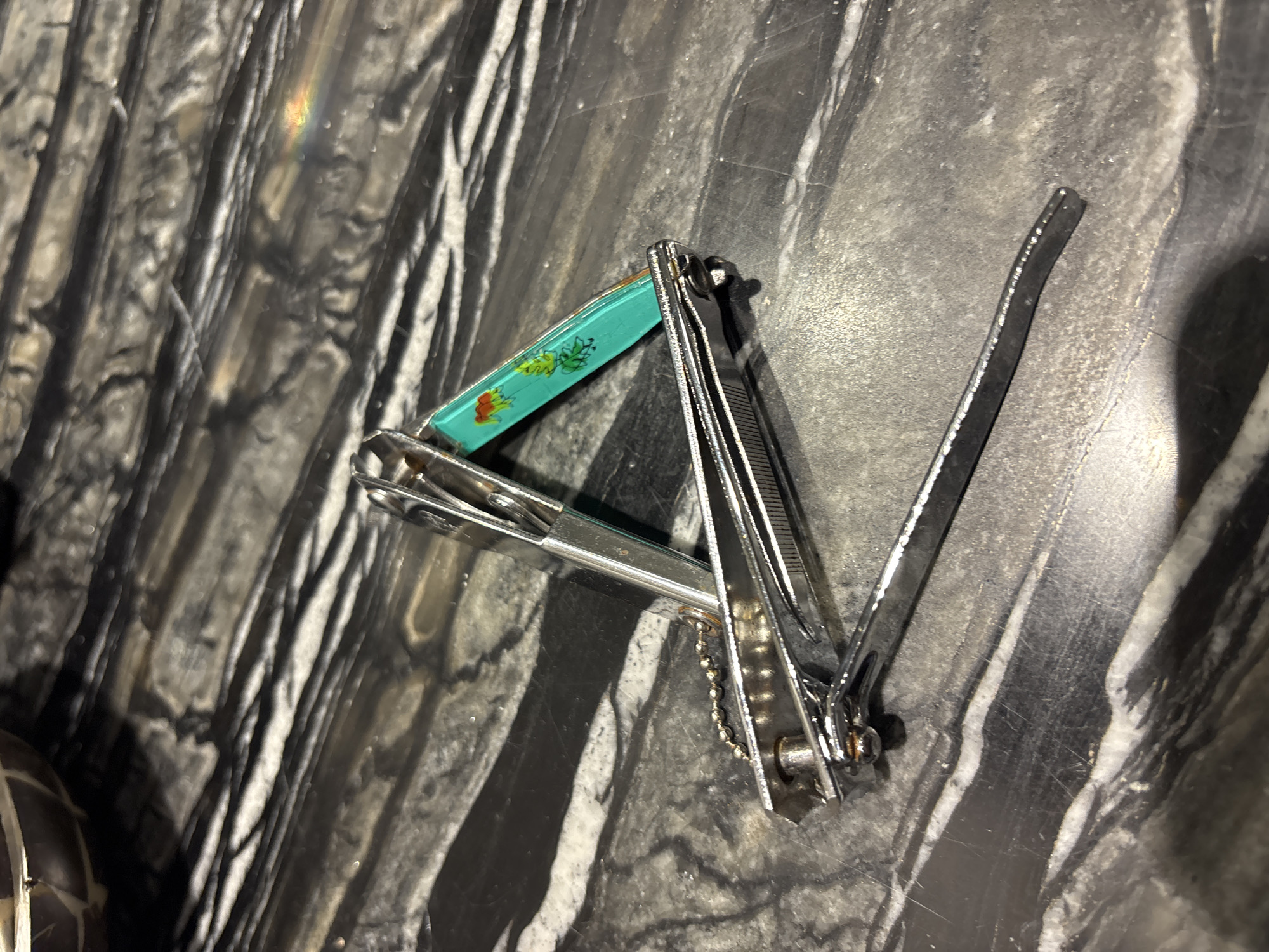 A pair of nail clippers rests on a polished, dark marble surface. The clippers are made of silver-toned metal and have a small, turquoise-colored handle with an orange and green floral pattern. The nail clippers are partially open, with the cutting blades facing upwards. The marble surface has prominent white and black veining, creating a dramatic, striated effect. The lighting in the image casts shadows from the nail clippers onto the marble. The scene is devoid of any human presence. The exact location within Dubai, United Arab Emirates, cannot be determined from the provided image.
