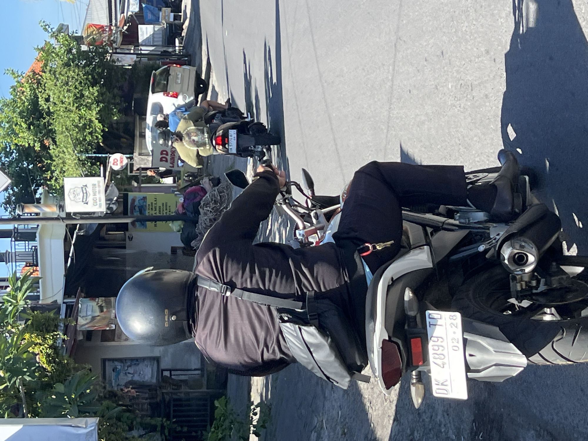 A back view of a person wearing a black helmet and black long-sleeved shirt is riding a motorcycle on a street in Sukawati, Indonesia. The motorcycle has a white license plate that reads "DK 4899 TD" and a smaller plate below that reads "02.28". A black messenger bag is slung across the rider's chest. The rider's legs are clad in black pants and dark dress shoes. In the background, other motorcycles and a white car are visible, along with buildings and lush green trees. The scene appears to be a sunny day, casting shadows on the road. Signs for "CUCI MOTOR" (motorcycle wash) and "DD LAUNDRY" are visible in the background.