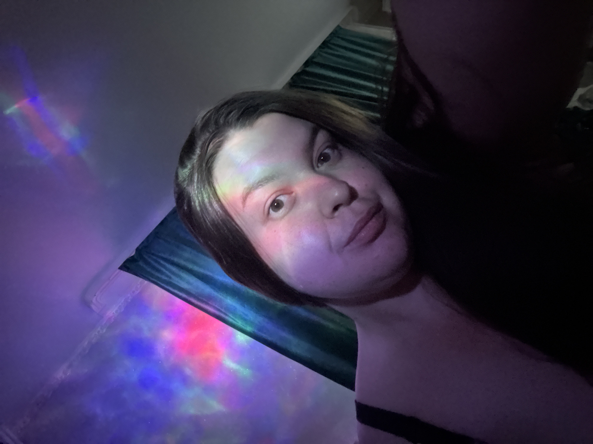 A selfie-style photograph captures a person with fair skin, dark hair, and a neutral expression. Light from a projector or similar source casts colorful patterns across their face and the surrounding surfaces. The person is wearing what appears to be a black top. A curtain is draped behind them. The location of the image is Марковский, Russia, according to the prompt.