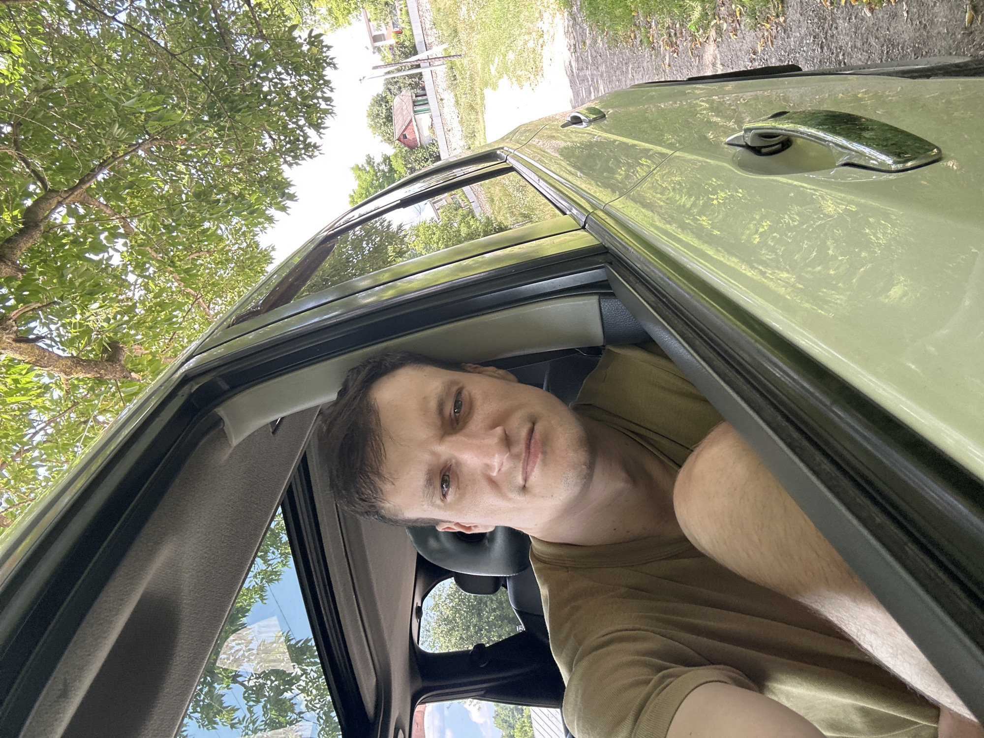 A man is taking a selfie from the driver's seat of a light green car. The car's sunroof is open, revealing green trees and a bright blue sky overhead. The man is wearing a tan t-shirt and his left arm and knee are visible. The car appears to be parked on a dirt road next to a grassy area with some fallen leaves. In the background, there are houses and fences, consistent with a rural setting in Makiv, Ukraine. The overall scene suggests a sunny day.