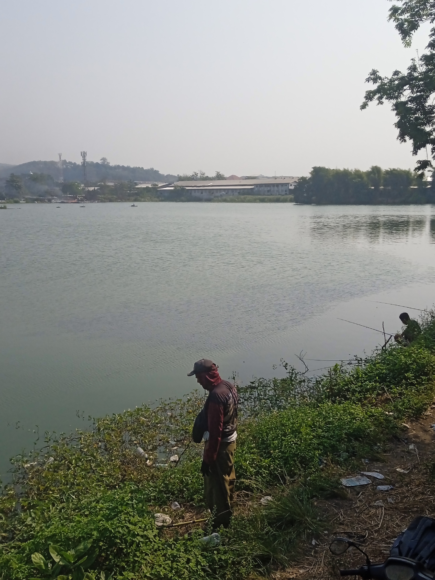 In Semarang, Indonesia, two individuals are observed fishing by a tranquil lake. The person in the foreground, clad in a red and black long-sleeved shirt, brown pants, and a dark baseball cap, stands facing away from the camera, his gaze directed towards the water. Behind him, another fisherman, wearing a dark green shirt and pants, is seated on the bank, also engaged in fishing. The lake's surface is calm, reflecting the overcast sky. The shoreline is overgrown with lush green vegetation, interspersed with scattered plastic debris. In the background, a cluster of buildings and communication towers are visible, hinting at the urban landscape surrounding the serene fishing spot.